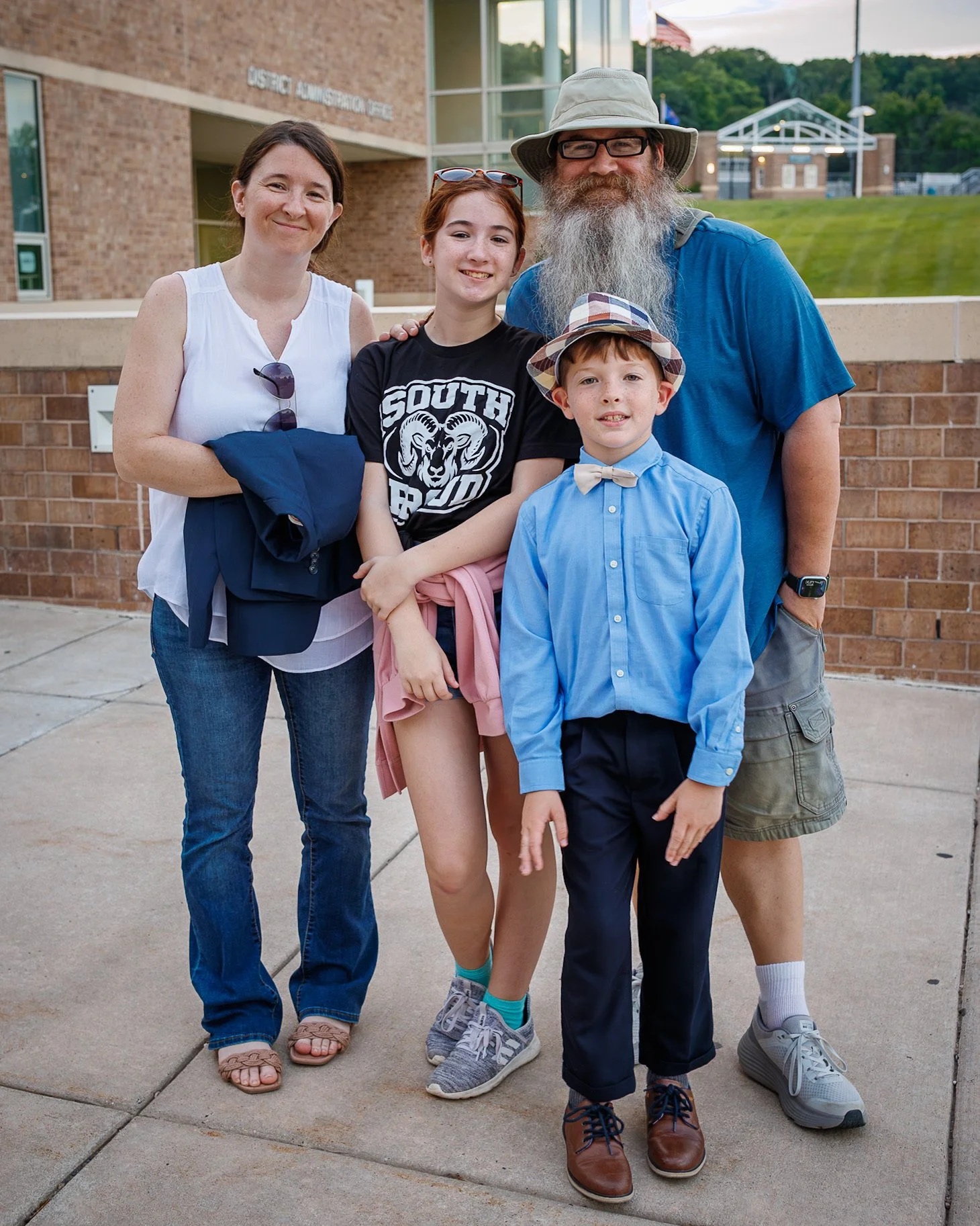 A family of five standing together outside a school building, smiling at the camera. The group includes a woman, a teenage girl, a man with a long beard, a young boy with a hat, and a girl with minimal clothing, suggesting warm weather.