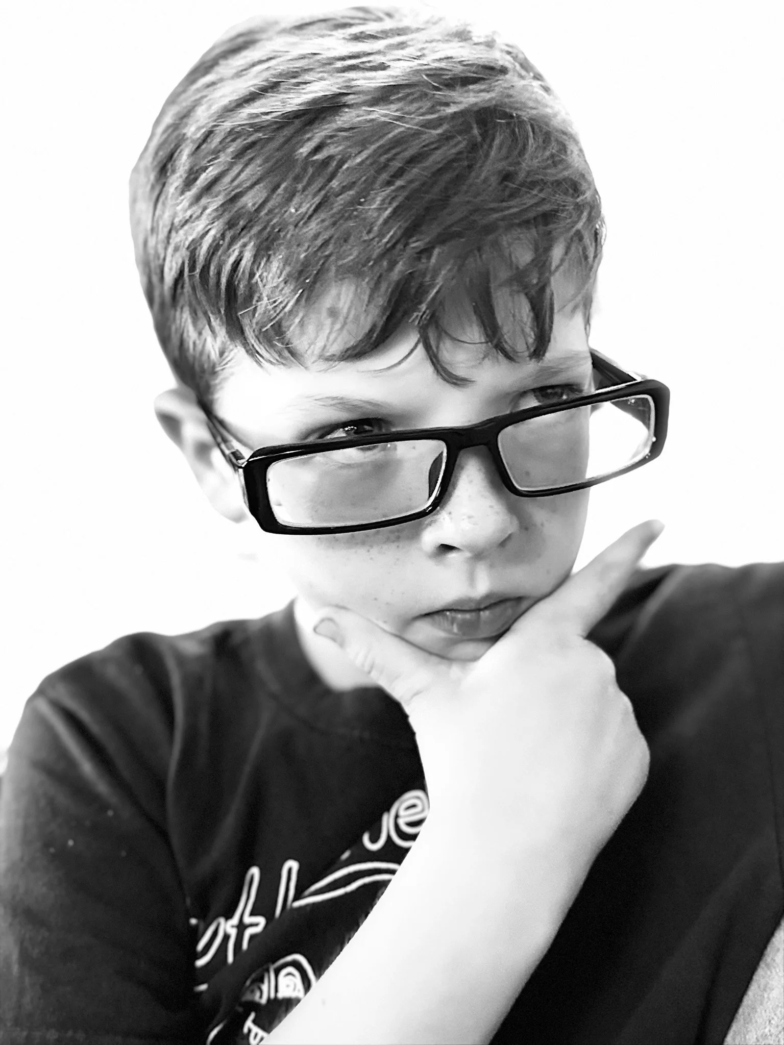 A black and white photo of a young person with short hair, wearing glasses and a dark shirt, with a hand resting near their cheek, looking thoughtful.