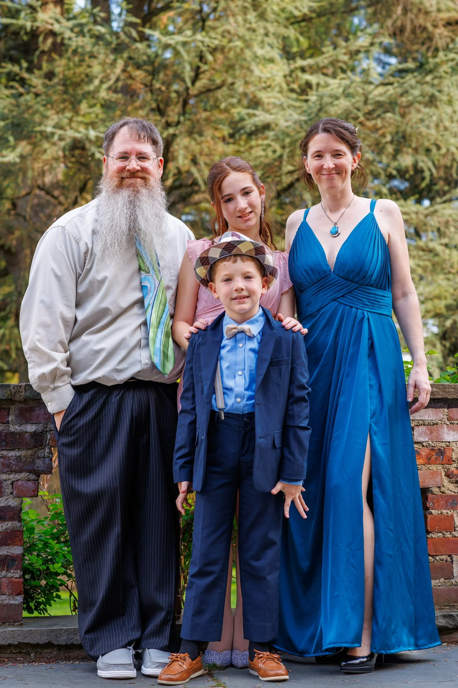 A family of five standing outdoors in front of green trees, smiling for a photo. The group includes an older man with a white beard and glasses, a young girl, a young boy in a tuxedo and bow tie, and two women, one in a blue dress and the other partially visible.