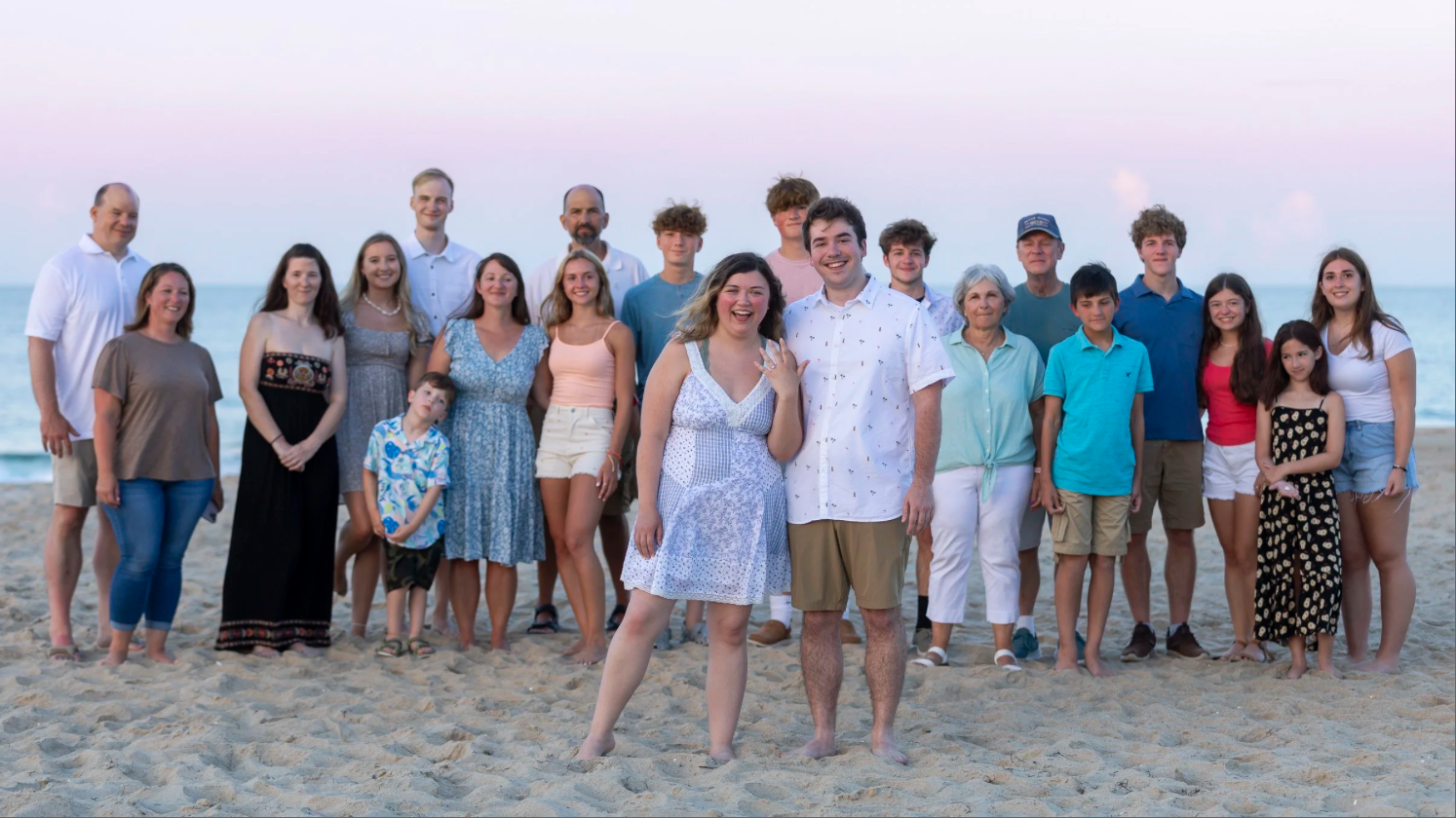 A group of 21 people, including children and adults, standing on a sandy beach with the ocean and a pastel-colored sky in the background, smiling and posing for a photo.