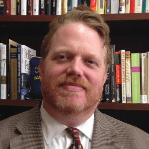 NIck Little looking straight at camera, bookcase in background, wearing brown suit and brown tie