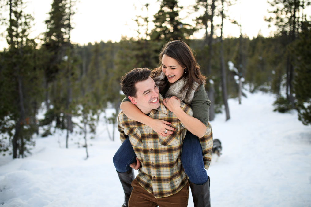 Smiling man giving woman a piggy back ride in snowy forest.