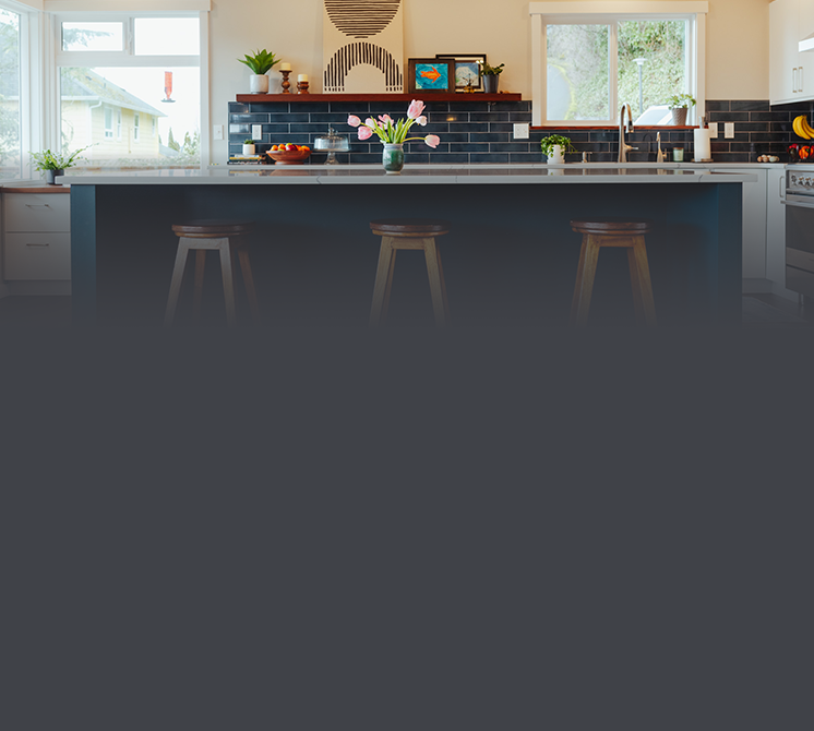 Modern kitchen with black subway tile backsplash, white countertops, and three wooden bar stools at a kitchen island. Decor includes a vase with pink tulips, potted plants, and framed artwork, with windows providing natural light.