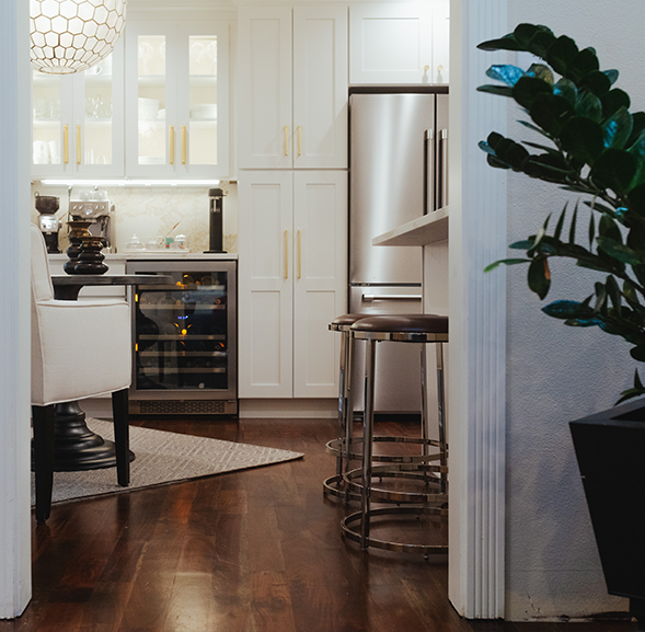 View inside a modern kitchen showing white cabinets with gold handles, a wine fridge, a small dining table with beige chairs, and a bar area with high stools, hardwood flooring, and a large green plant.