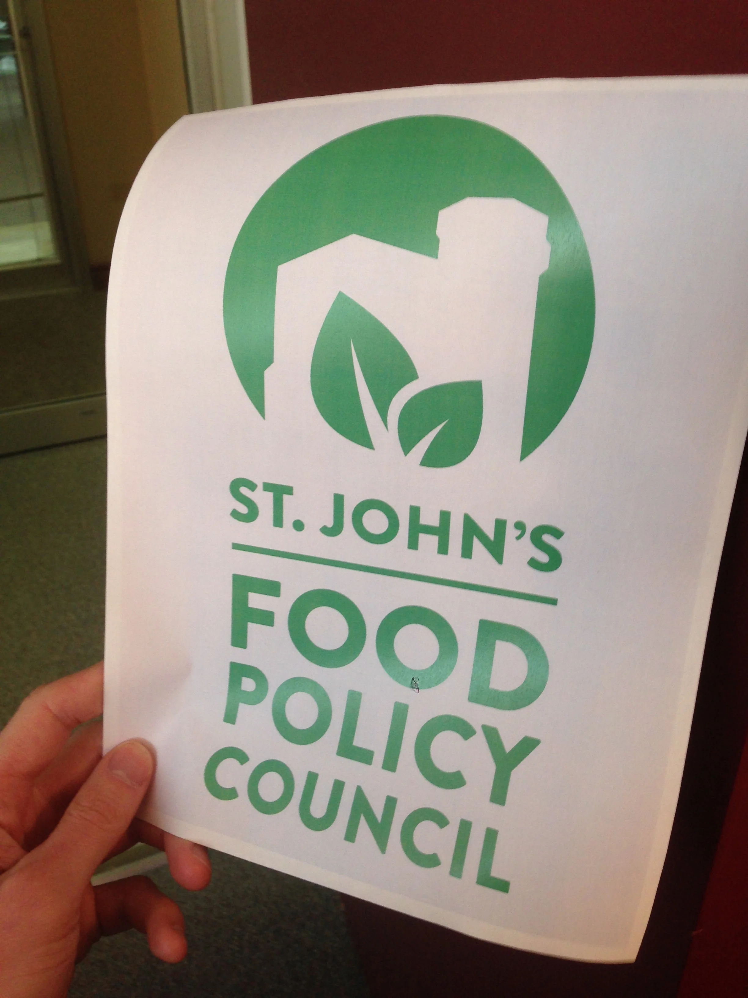 A hand holding a handout featuring the St. John's Food Policy Council title and logo in green. The logo is a sillhoute of Cabot Tower with two green leaves inside.