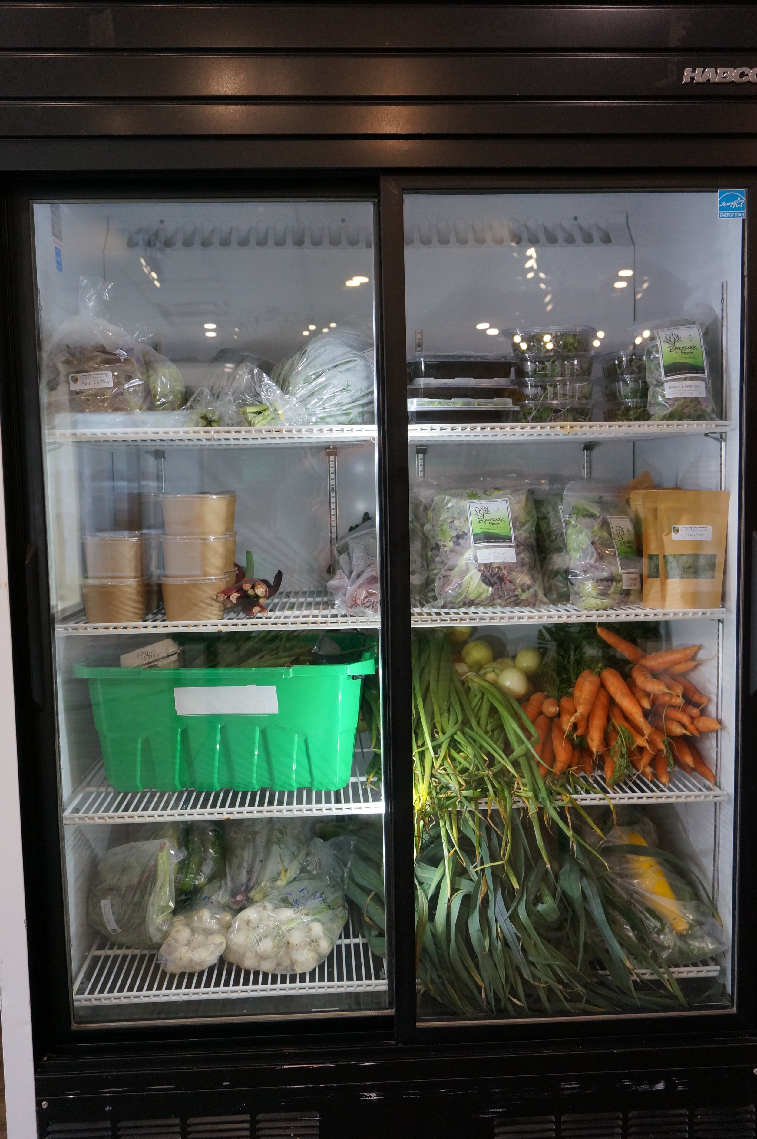  A commercial cooler fridge stocked with local greens, carrots, onions, spring onions, zucchini, eggs, and small containers of food. 