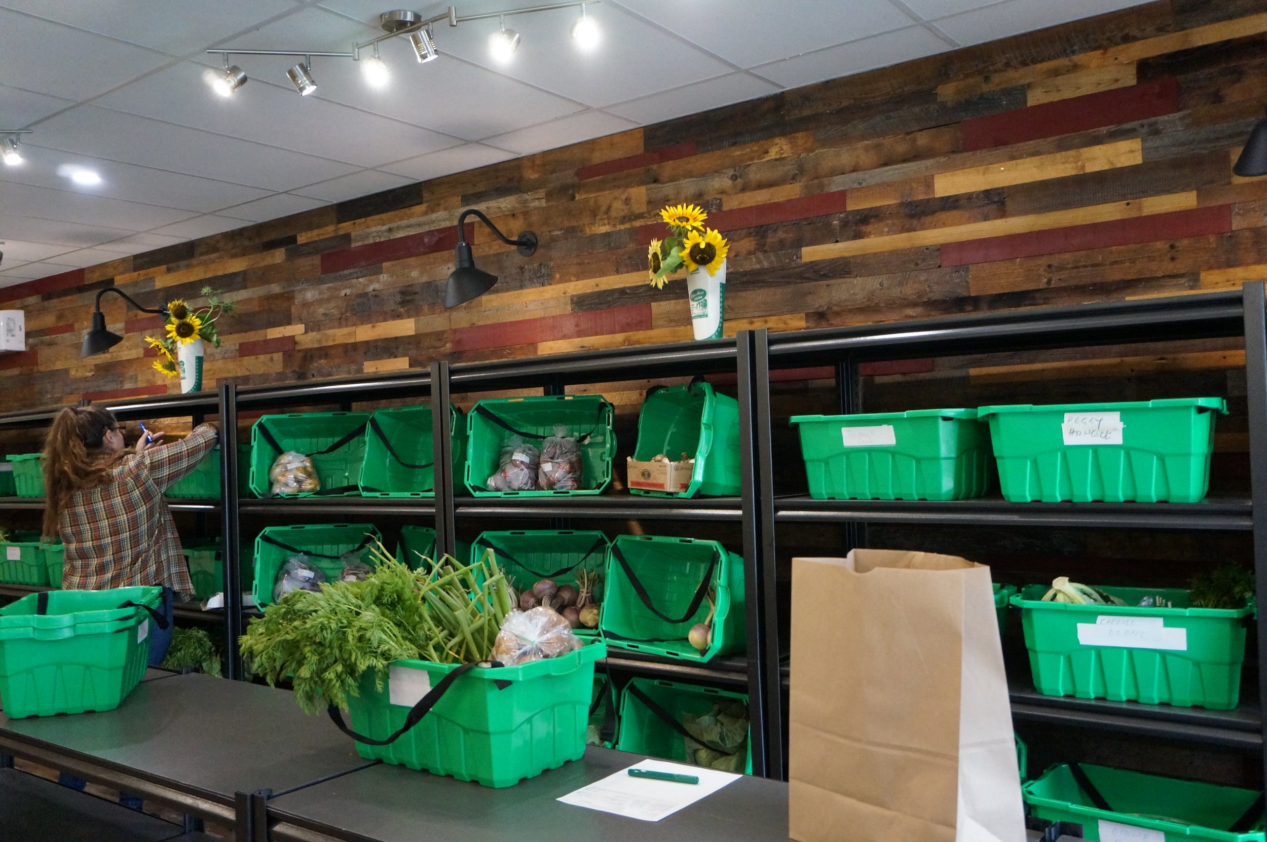  A wall of heavy duty shelving against a wood paneled wall. Each shelf holds two green plastic bins, each a different customer order. One bin is sitting on a counter in front of the shelves, a large paper bag next to it. 
