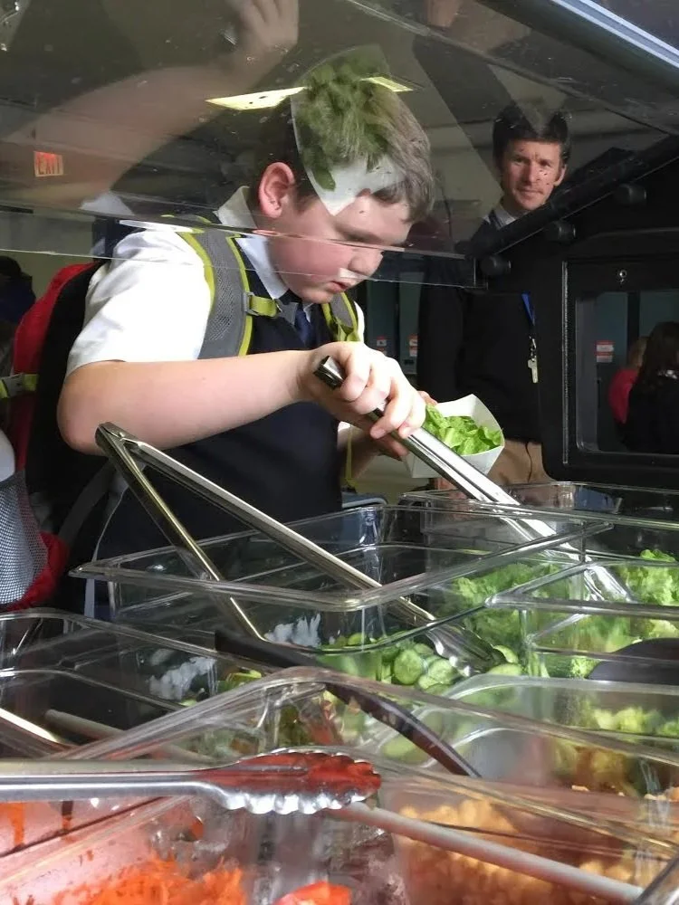 A young boy wearing a school uniform and backpack carefully selecting fresh salad greens from a salad buffet.