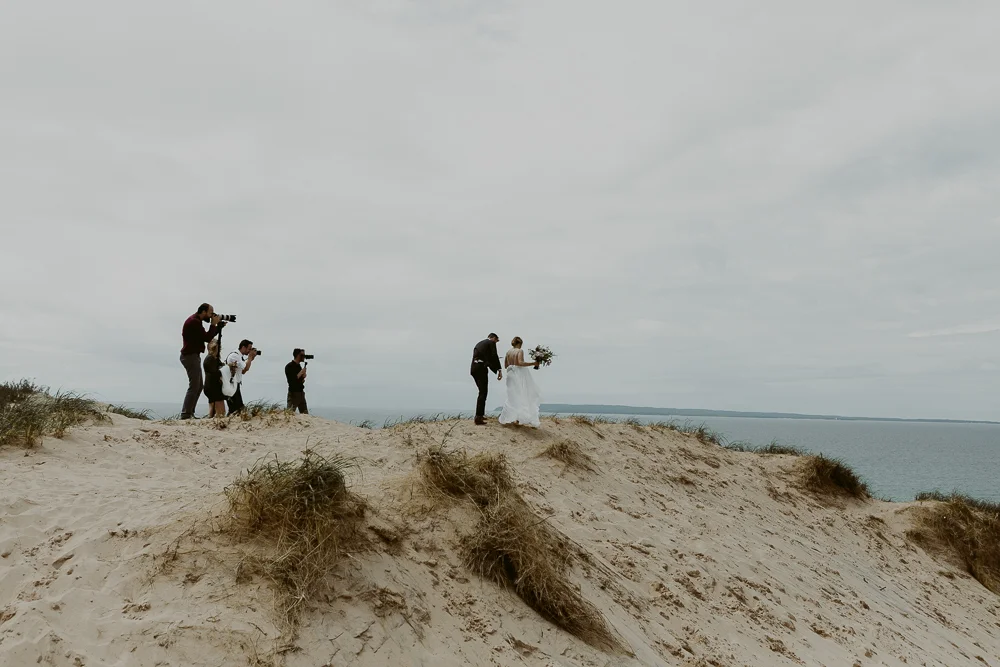 A wedding ceremony on a sandy beach with a bride holding a bouquet and a groom standing beside her, surrounded by photographers capturing the moment.