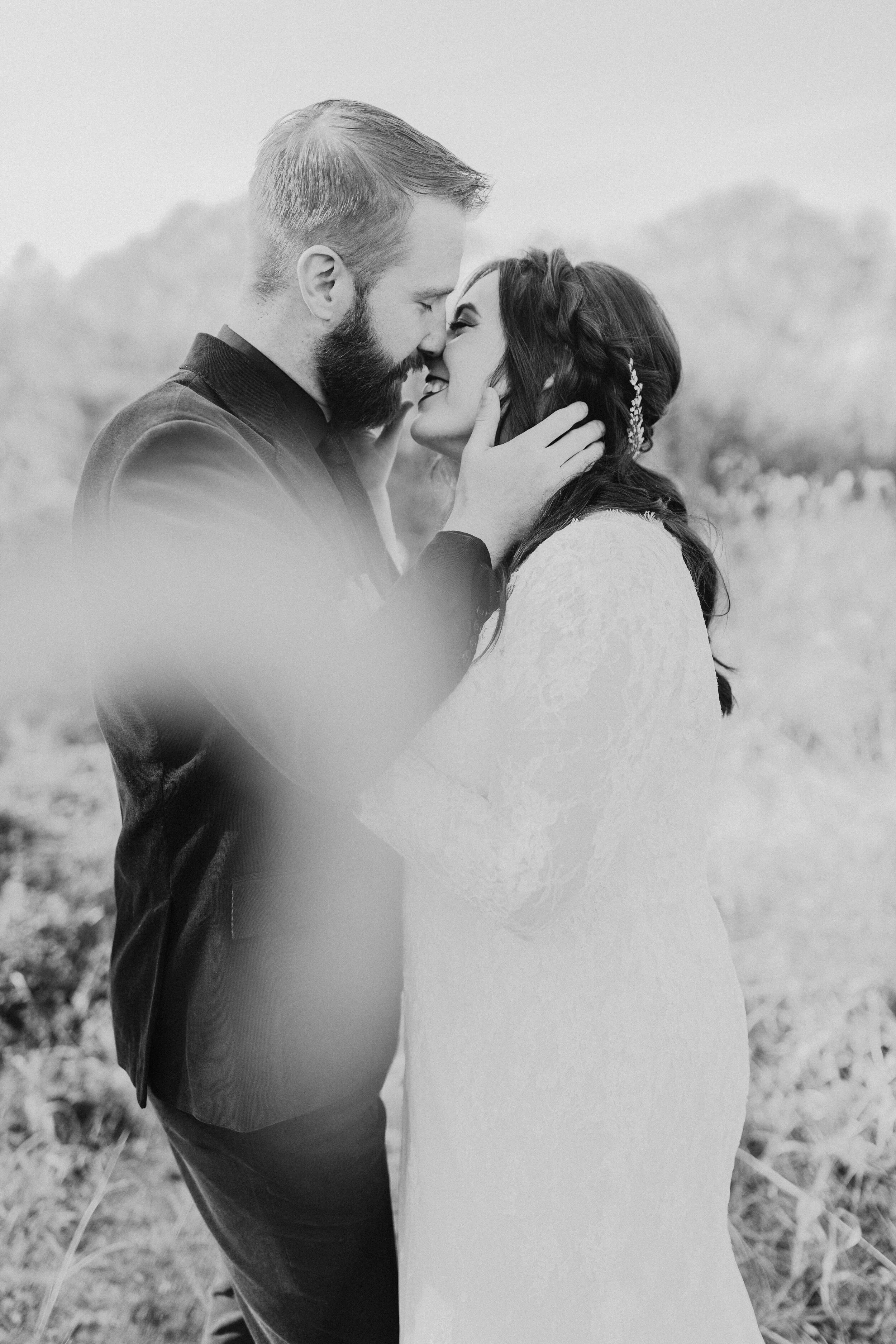 A black-and-white photo of a couple embracing closely, about to kiss, outdoors with a blurred natural background.