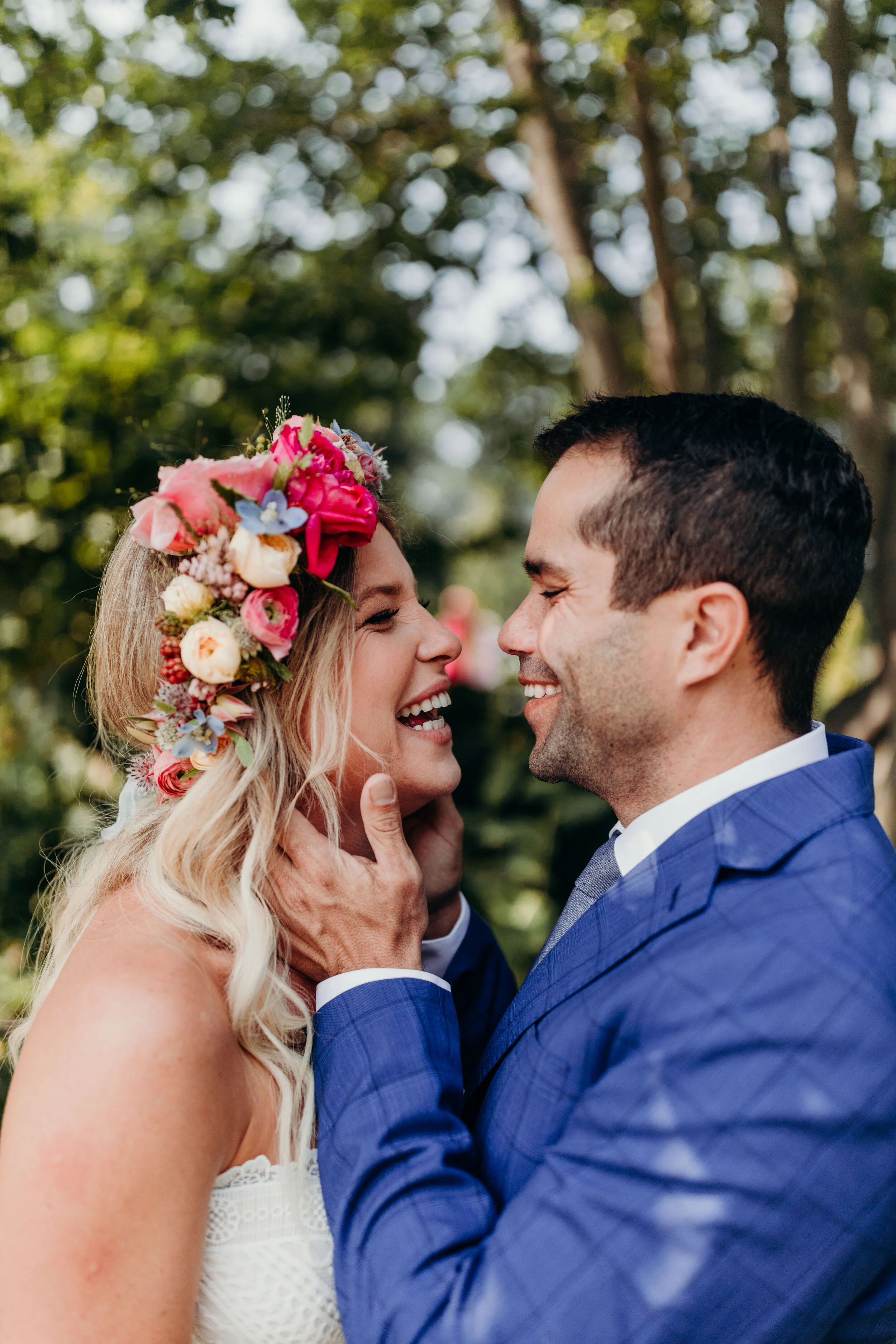 A wedding couple smiling and holding each other's faces in a forest setting, woman with a colorful flower crown and white dress, man in a blue suit and gray tie.