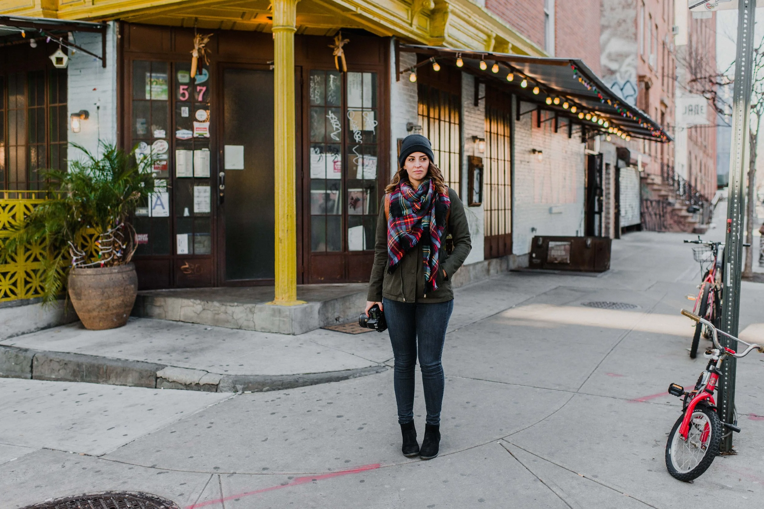 Woman standing on sidewalk near a building, holding a camera, wearing a black beanie, a colorful plaid scarf, dark green jacket, and black boots, in an urban street setting.