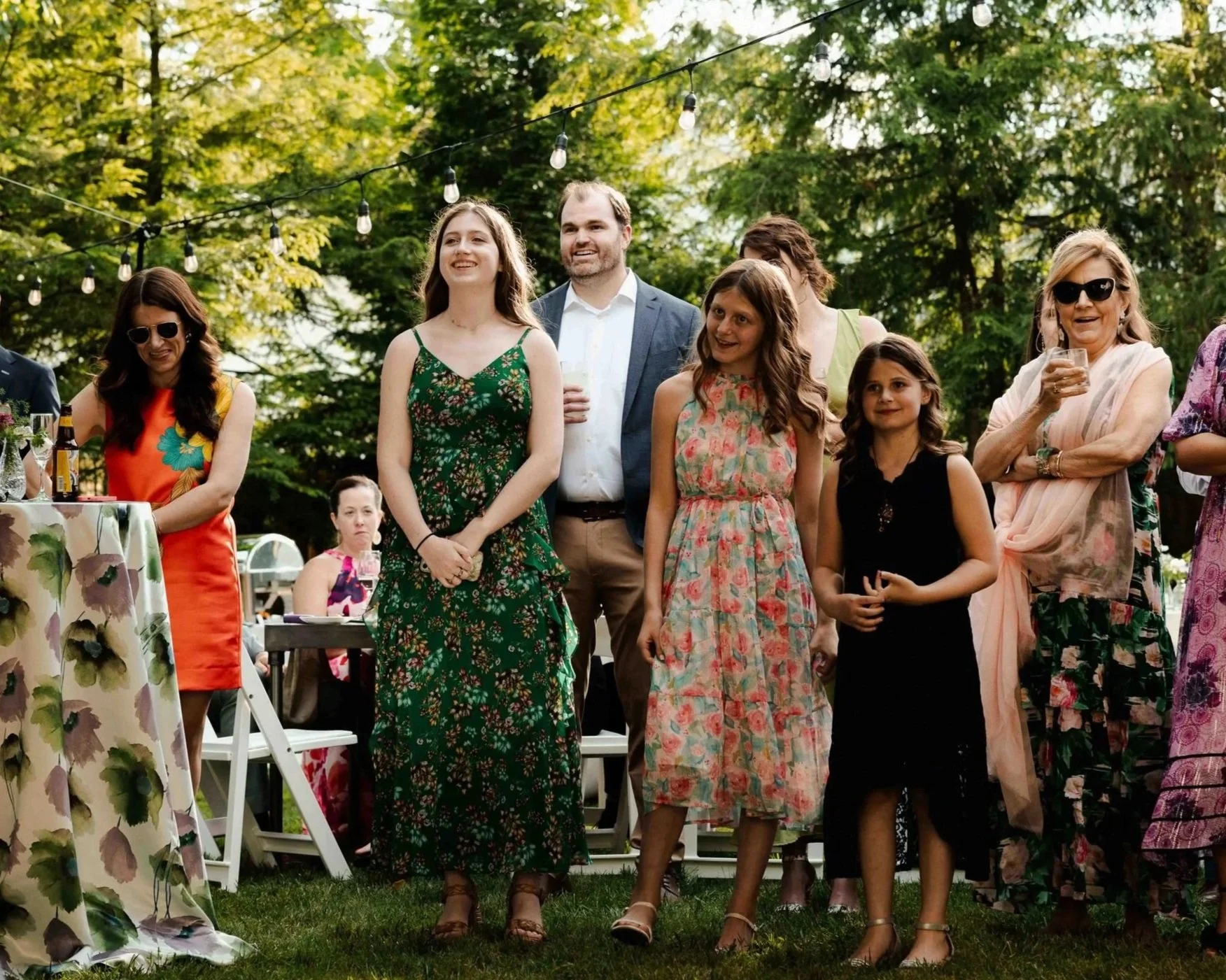 People enjoying an outdoor social gathering or party in a garden, with women and children dressed in colorful summer dresses, under string lights and surrounded by trees.