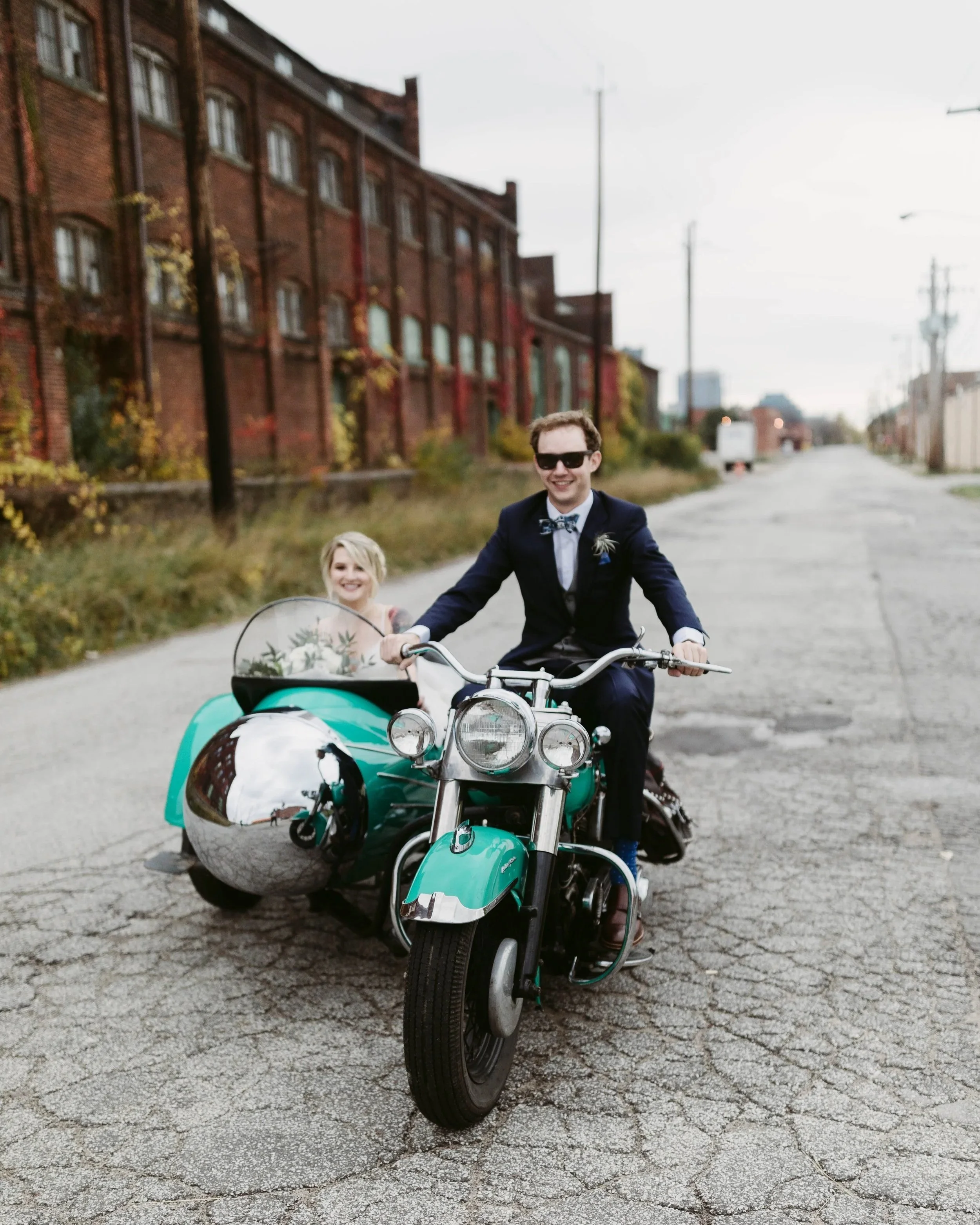 A smiling man in a suit and sunglasses rides a vintage turquoise motorcycle with a sidecar, with a woman in a white dress sitting in the sidecar on an empty city street with old brick buildings in the background.