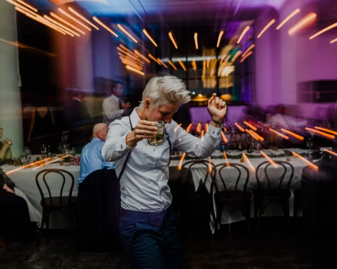 Woman dancing with a drink in a lively, brightly lit restaurant or club with colorful lights and empty tables in the background.