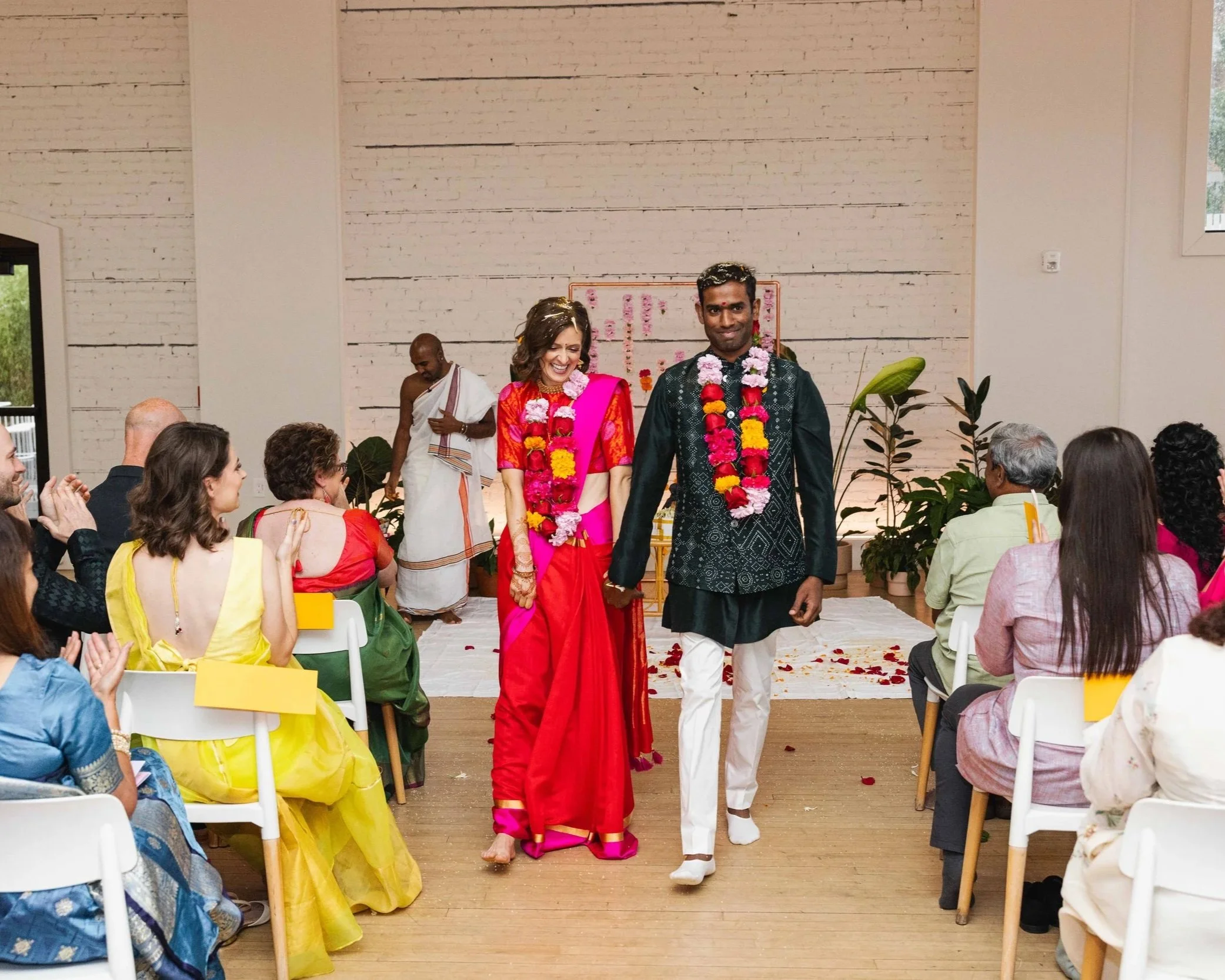 Couple wearing traditional Indian attire walking down the aisle during a wedding ceremony, surrounded by seated guests clapping and celebrating.