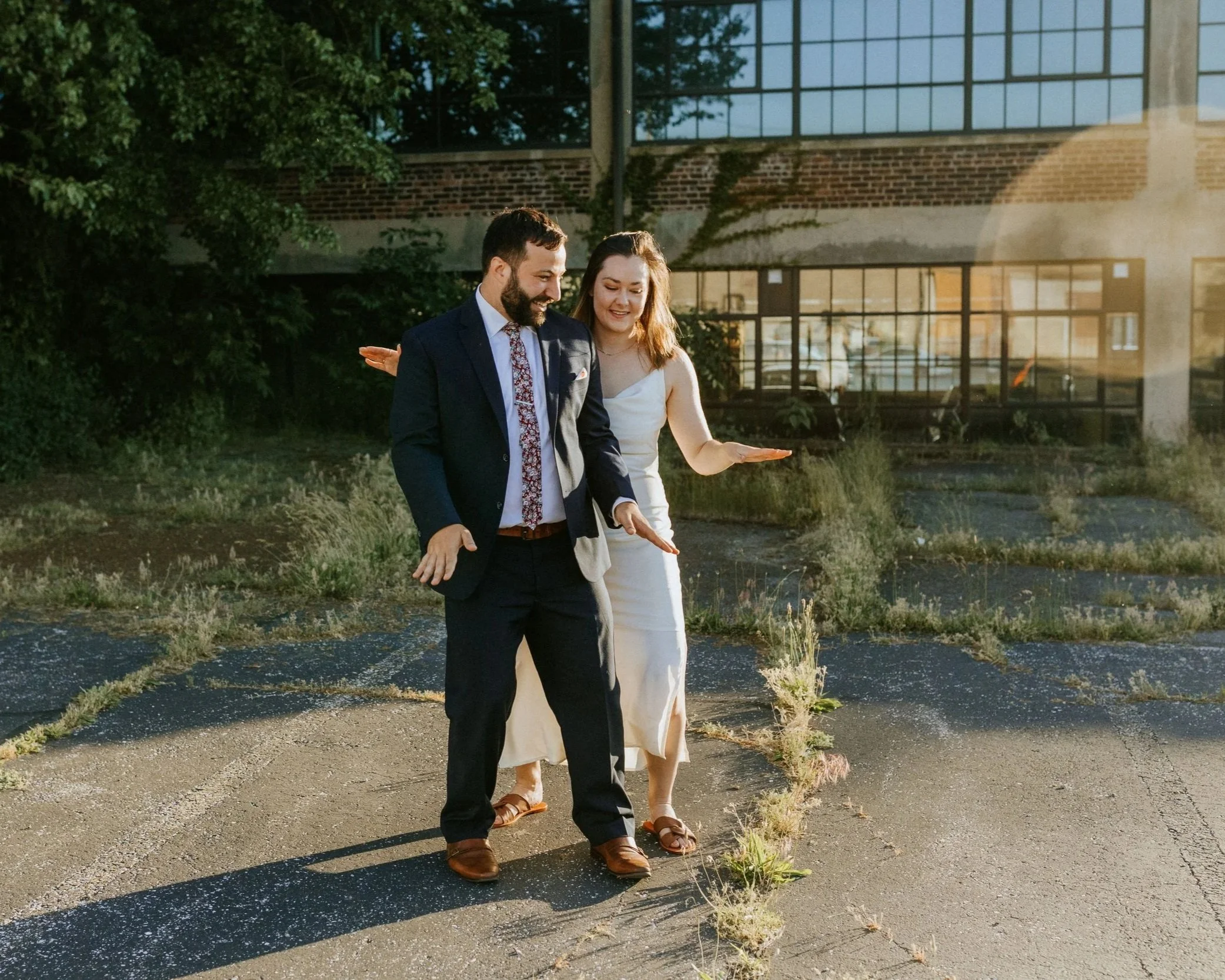 A man and woman walking outdoors on a sunny day, smiling and looking at the ground, with patches of grass growing through cracks in the pavement.