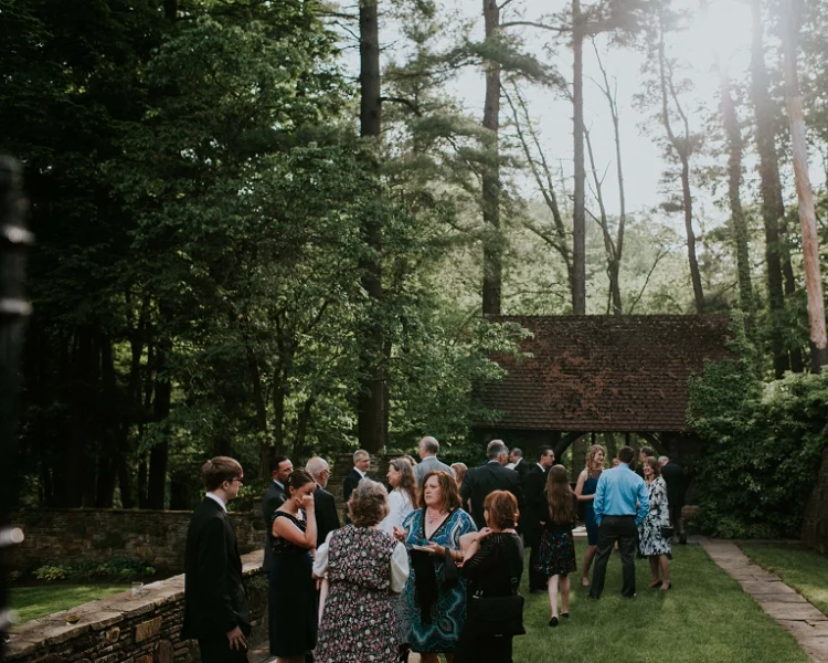 Group of people gathered outdoors in a lush, wooded area, attending an event or celebration in front of a small brick structure with a red-tiled roof.