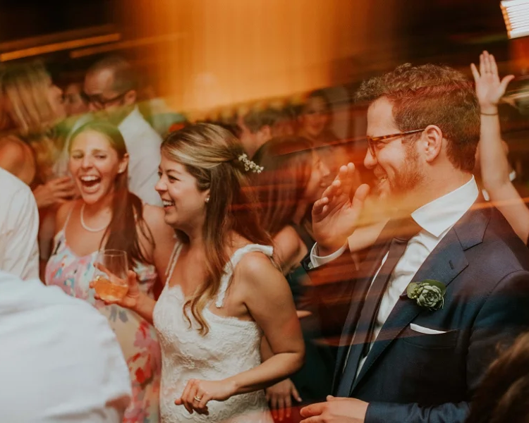 People dancing and celebrating at a wedding reception, including a bride in a white dress and a groom in a suit, smiling and enjoying the moment.