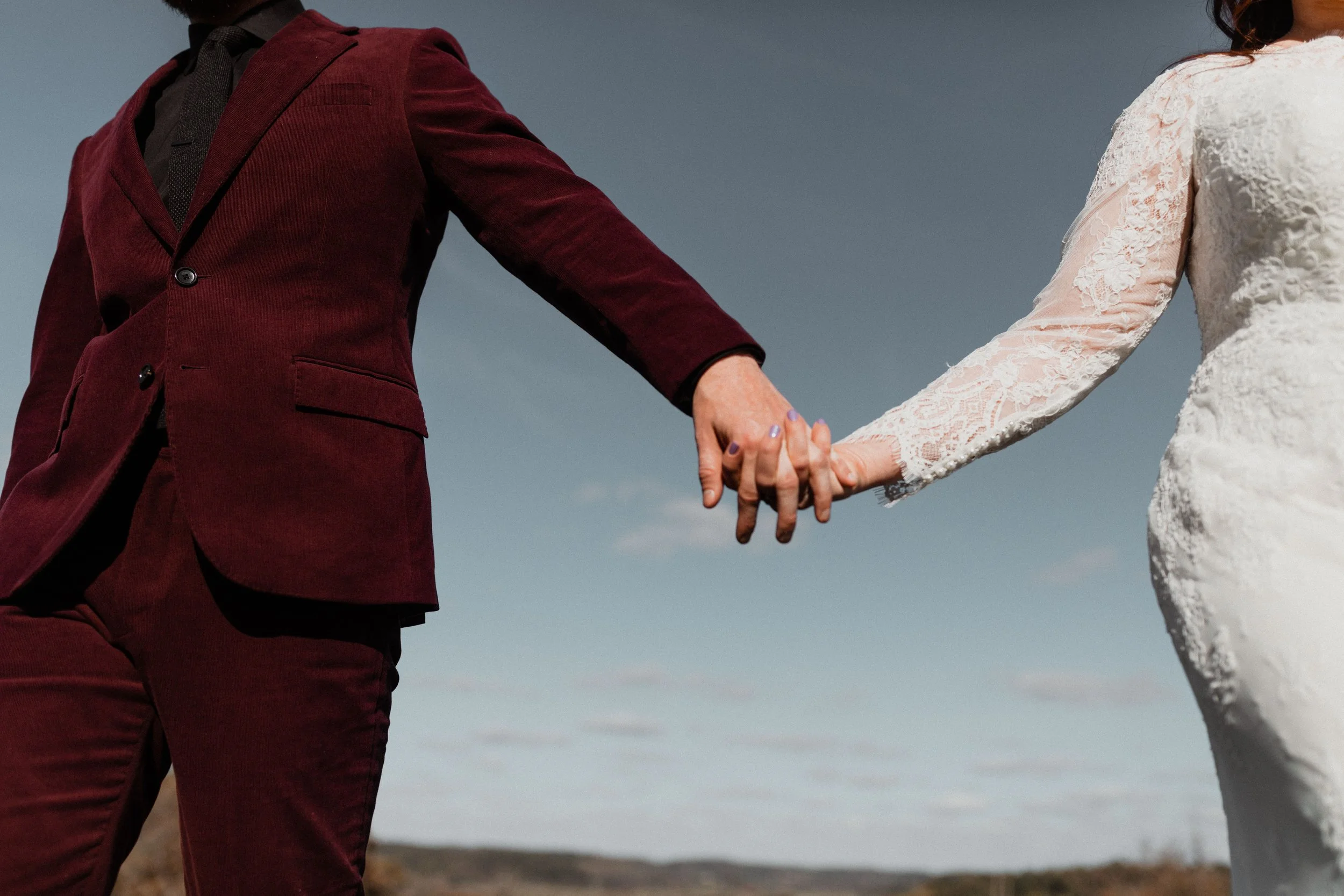 A man and woman holding hands outdoors, the woman wearing a white lace dress and the man in a dark red suit.