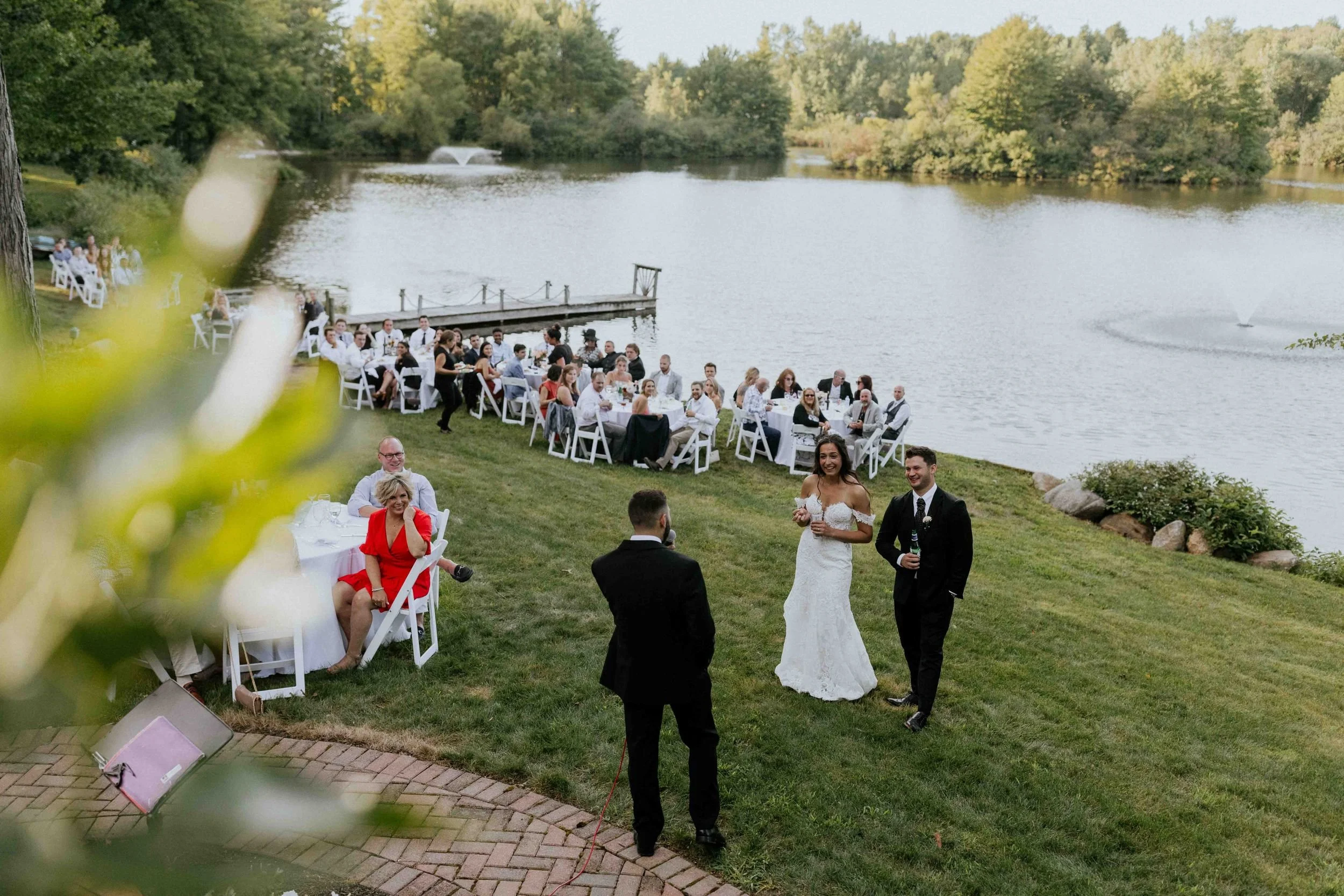 Outdoor wedding reception by a lake with guests seated at tables, two bridesmaids and a groom standing for photos, and a lake with trees in the background.