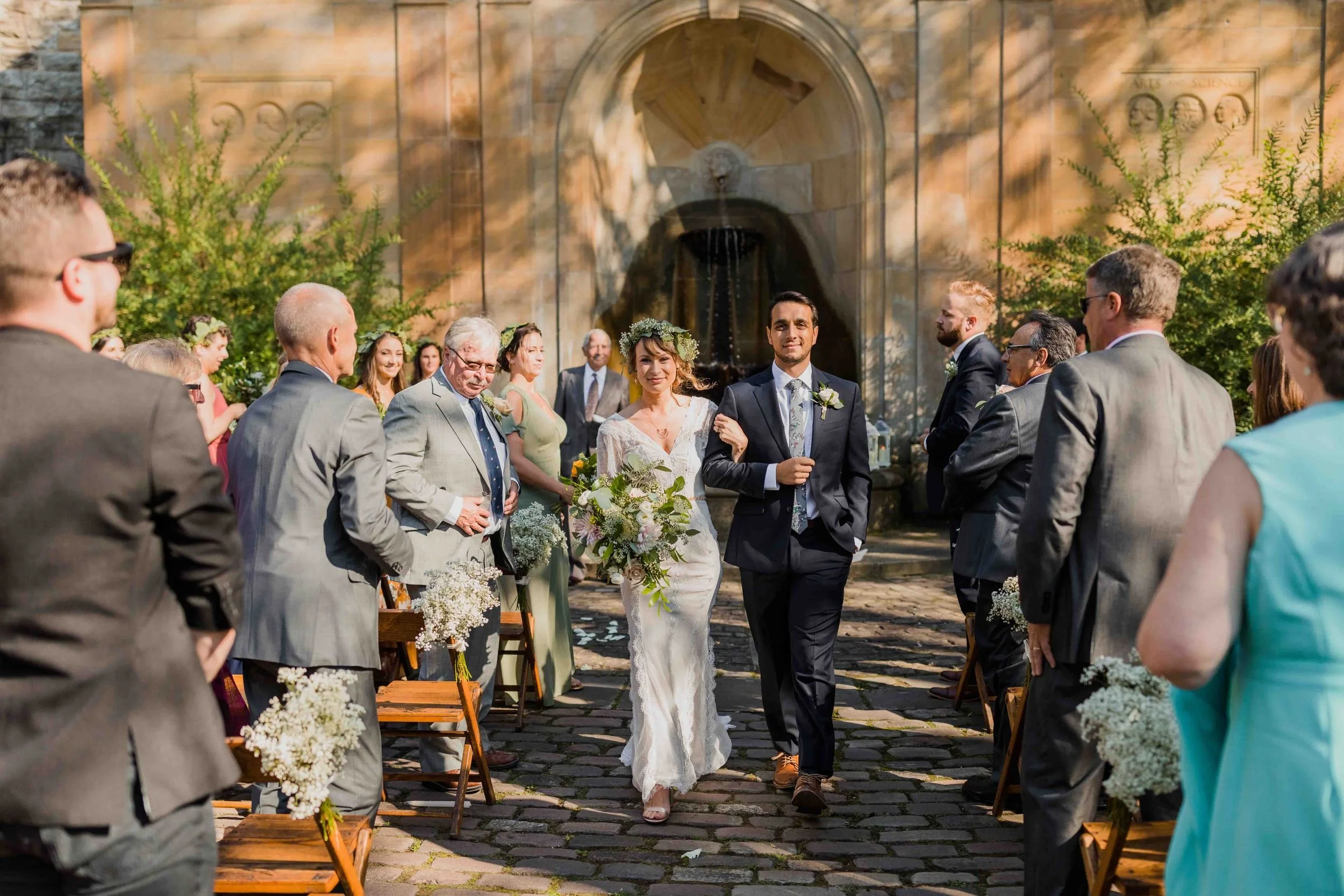 Bride and groom walking down the aisle after wedding ceremony outdoors, surrounded by guests seated on wooden chairs, with a stone fountain and lush greenery in the background.