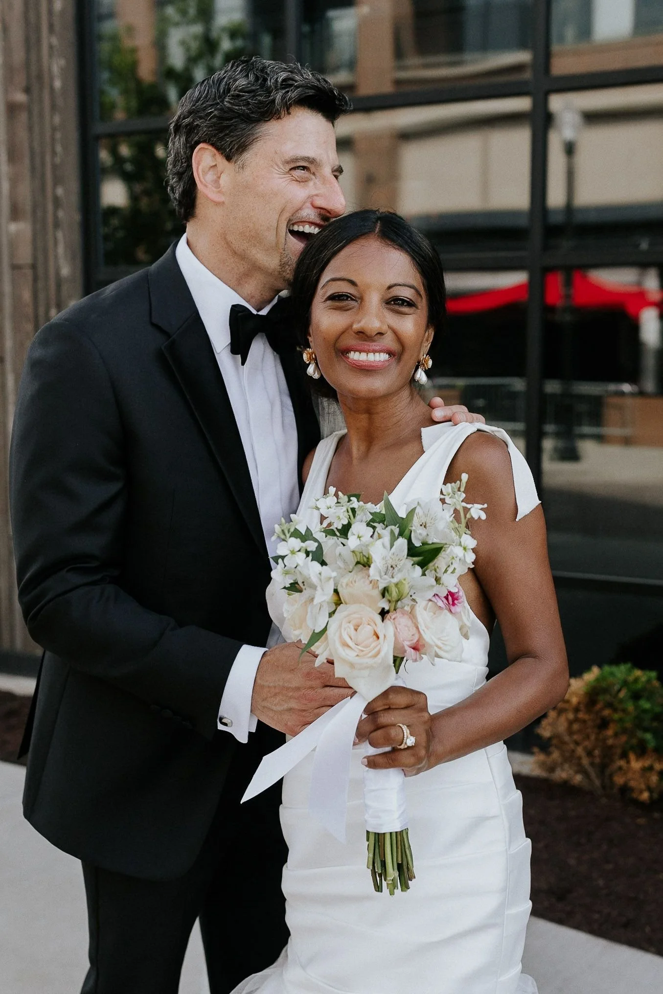 A newlywed couple, the groom in a black tuxedo and the bride in a white wedding dress, smiling and posing outdoors with the bride holding a bouquet of white and pink flowers.