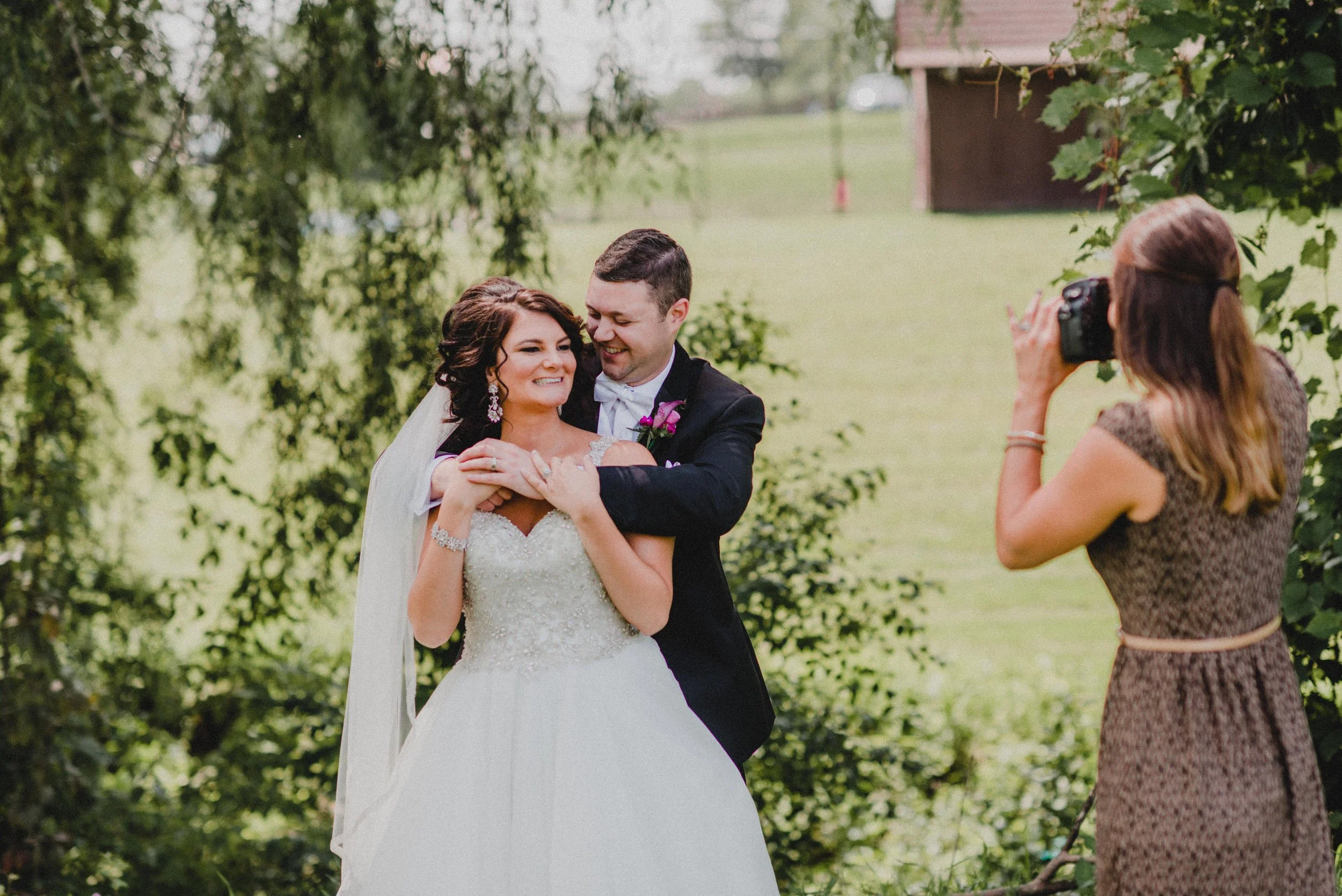 Bride and groom smiling and hugging during their wedding photo shoot outdoors, with a photographer capturing the moment.