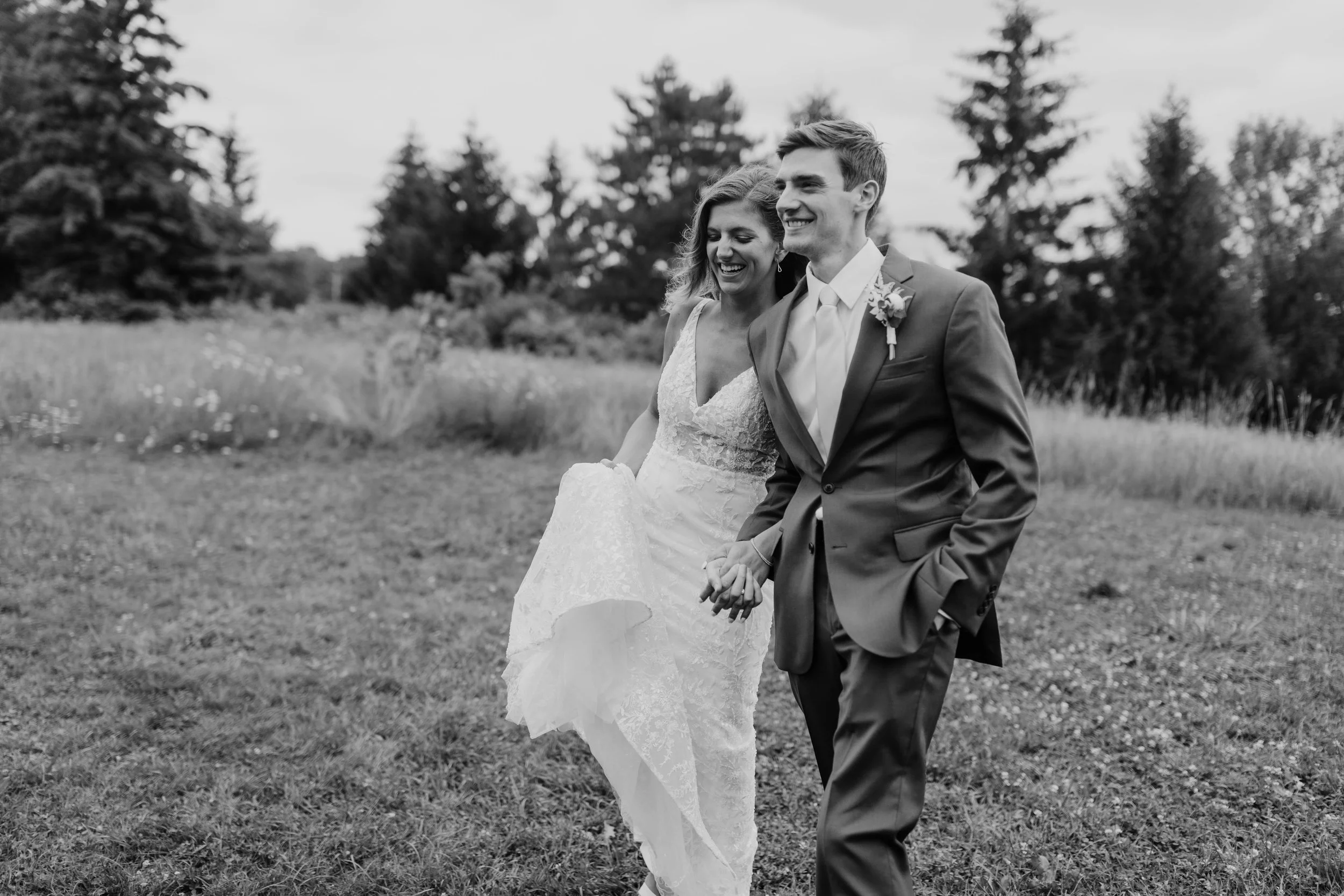 Black and white photo of a smiling bride and groom walking hand in hand in a grassy outdoor area with trees in the background.