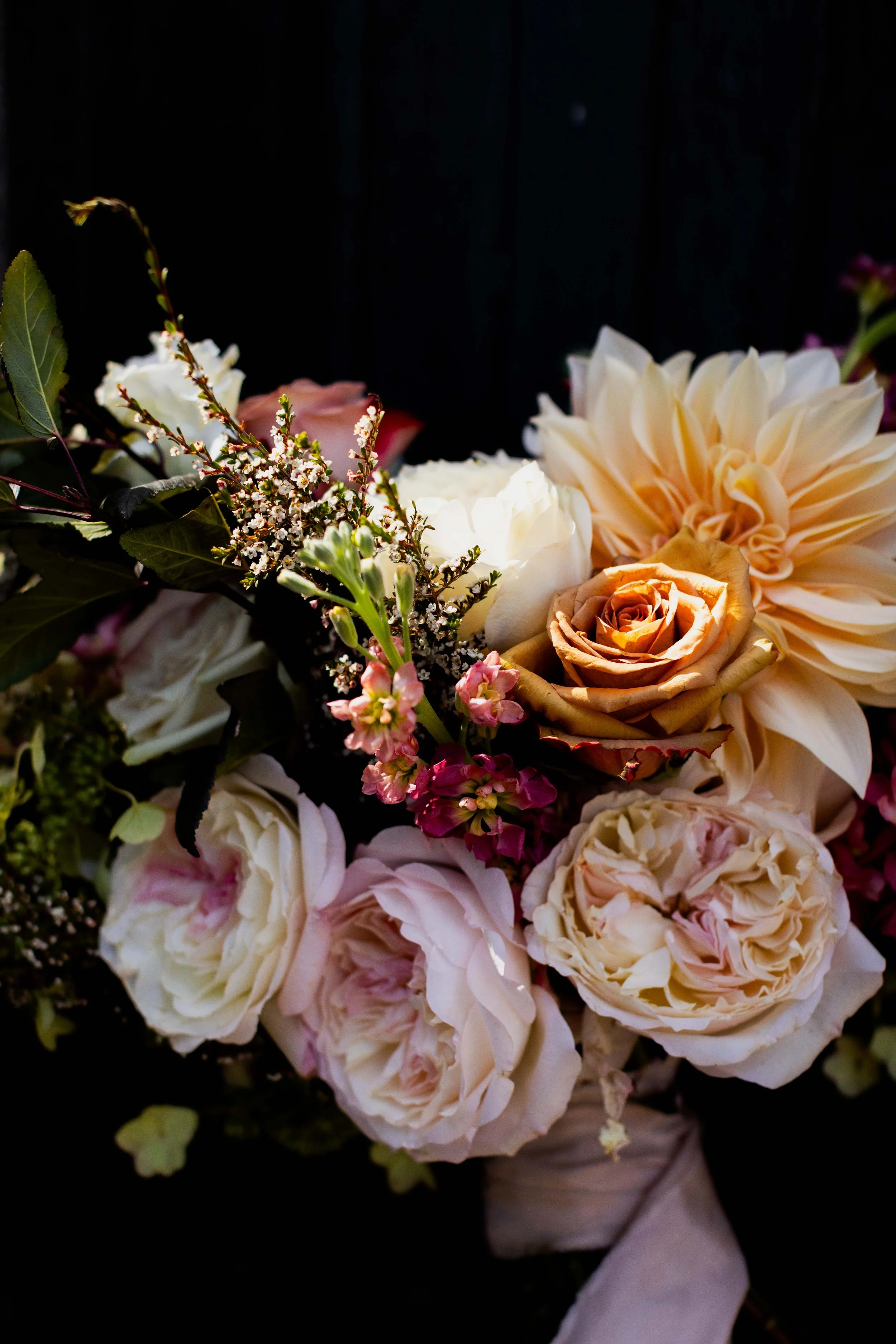 A bouquet of light pink, peach, white flowers including roses and dahlias with green leaves against a dark background.