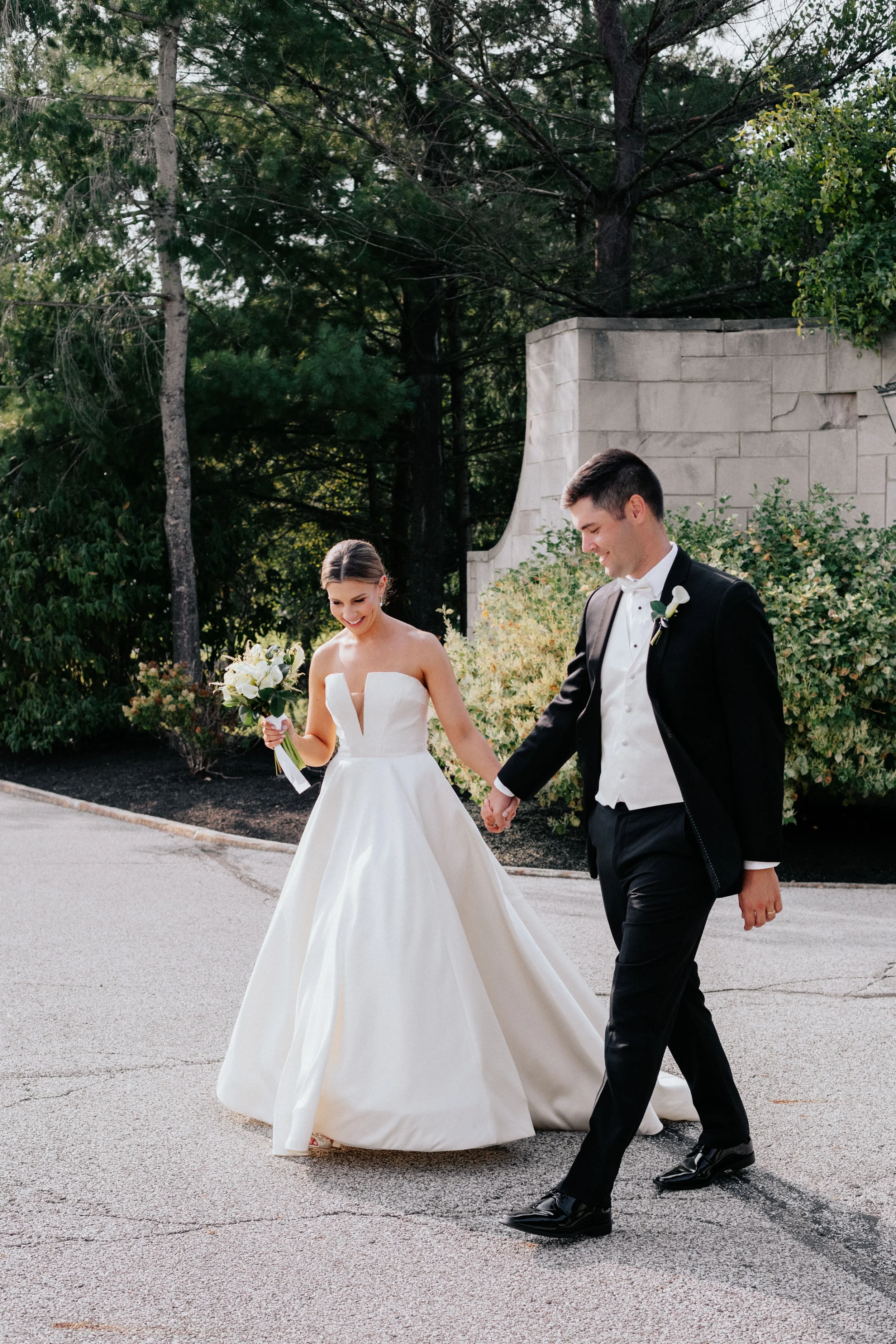 A bride and groom walking hand in hand outside, smiling, with the bride holding a bouquet of white flowers. The bride wears a strapless white wedding gown, and the groom is in a black tuxedo with a white shirt and vest. They are surrounded by greenery and trees.
