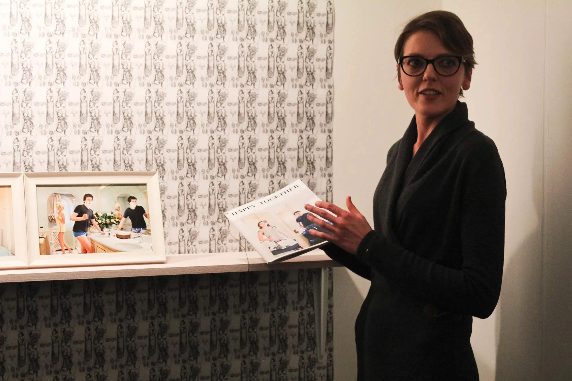 Francesca Catastini holding the magazine from of her series ‘Happy Together’ (2010), with framed prints from it on the shelf, and a repeating print of female genitalia as the wallpaper.