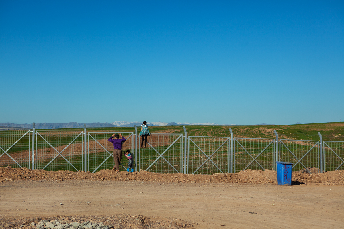 Siblings look through the perimeter fence of Darashakran Refugee camp to surrounding wheat fields. Erbil, Iraq. February 16, 2016. Photo by Danish Refugee Council/Eduardo Soteras Jalil.