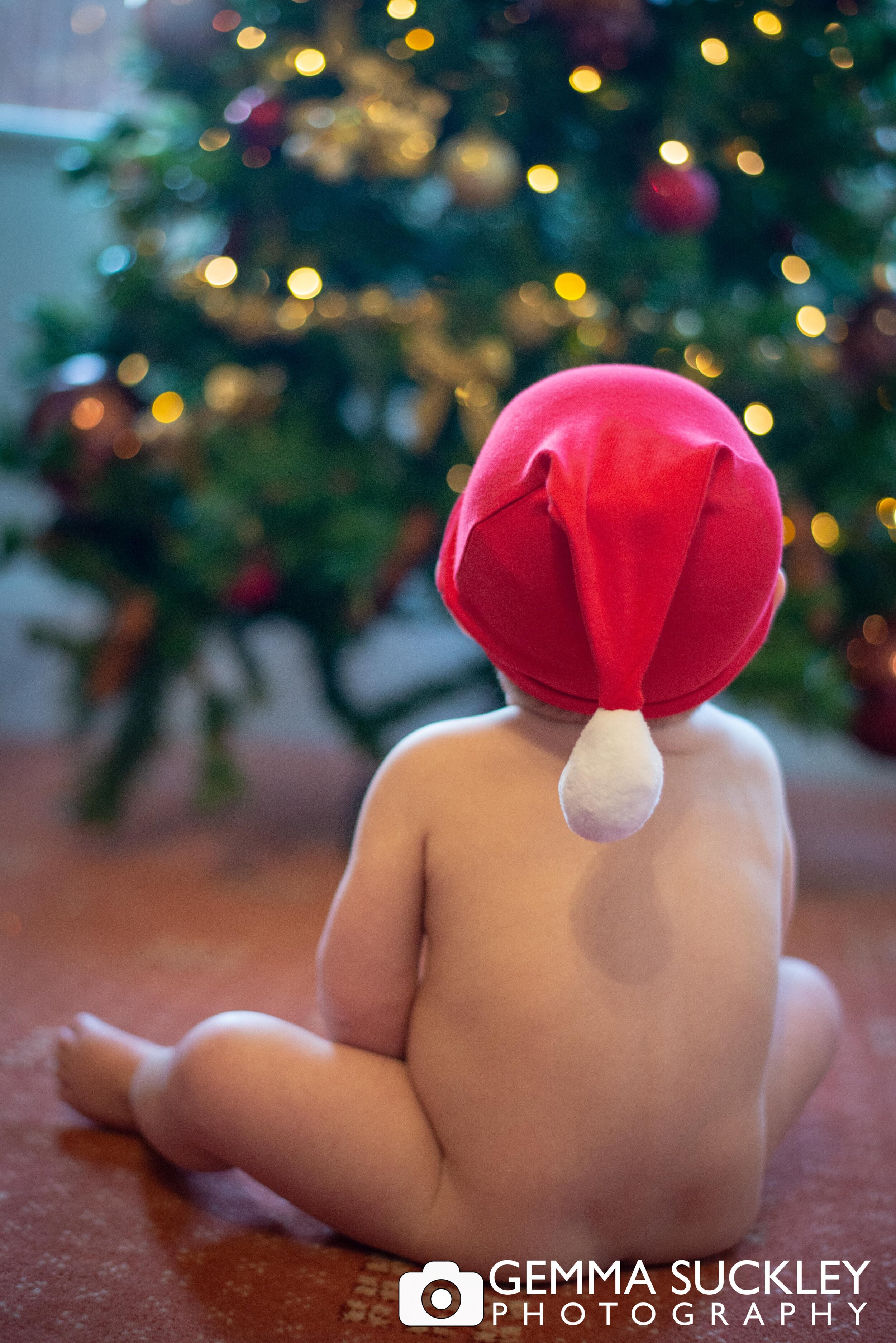 naked baby with santa hat looking at the christmas tree