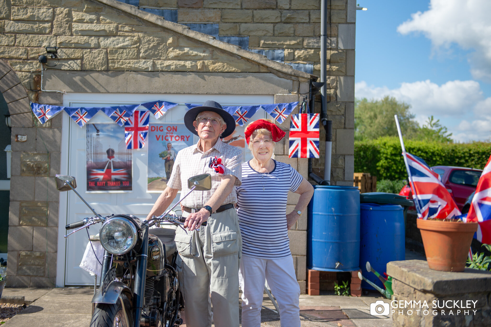 a doorstep portrait of an elderly man with his vintage motorbike and his wife looking proud on VE day