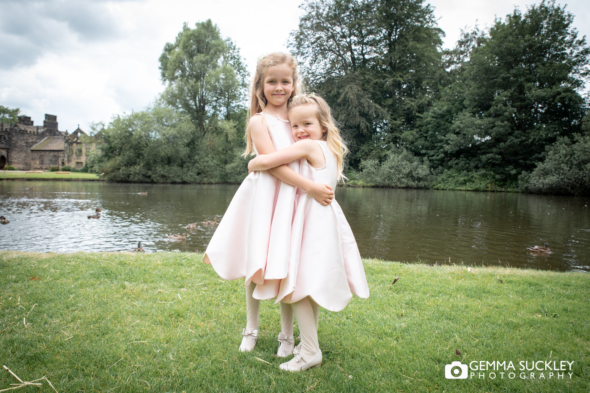 two flower girls smiling for the camera infront of the lake at east riddlesden hall