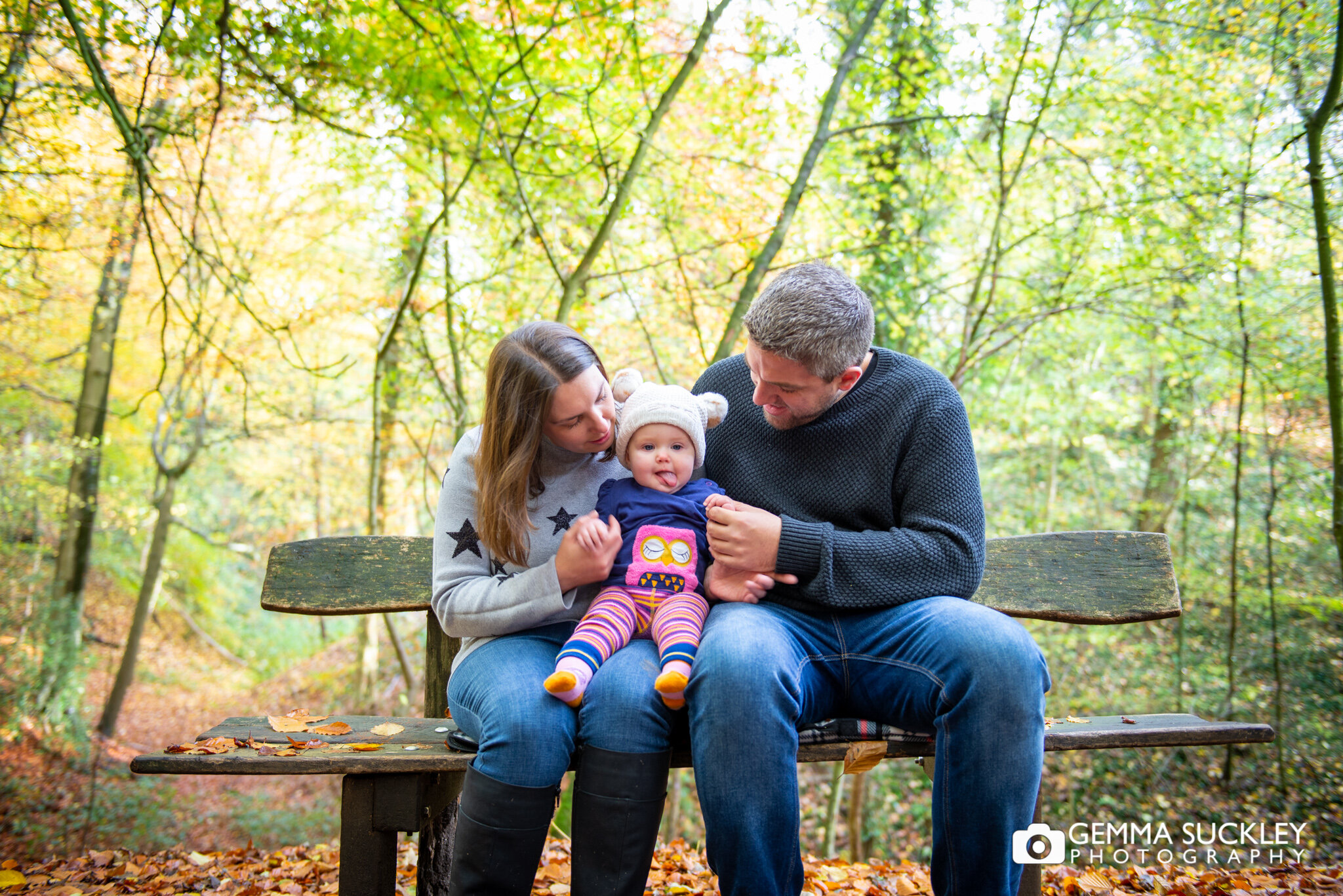 husband and wife sitting on a bench with their baby girl in skipton wood