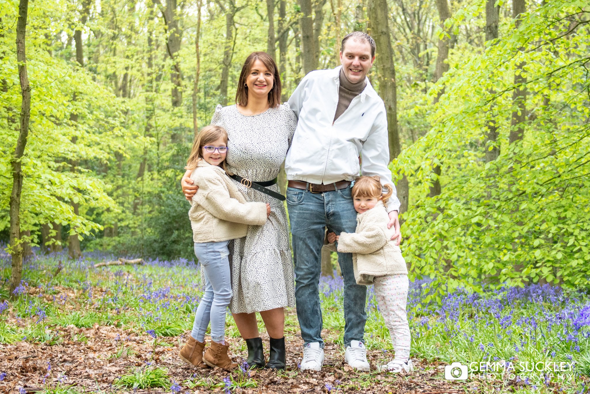 engaged couple with the two children hugging in the middle of a bluebell wood