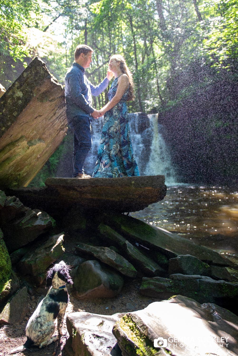 a couple hold hands in front of goitstock waterfall and their puppy looking up at them