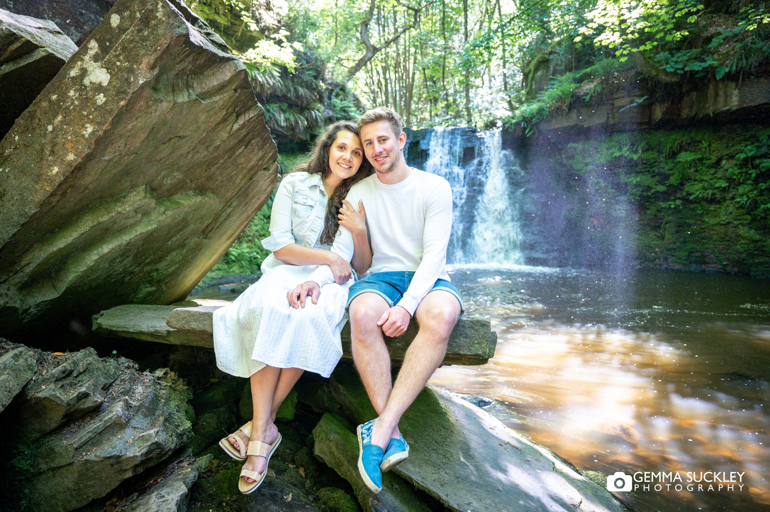 an engaged couple sitting on a rock in front of a waterfall