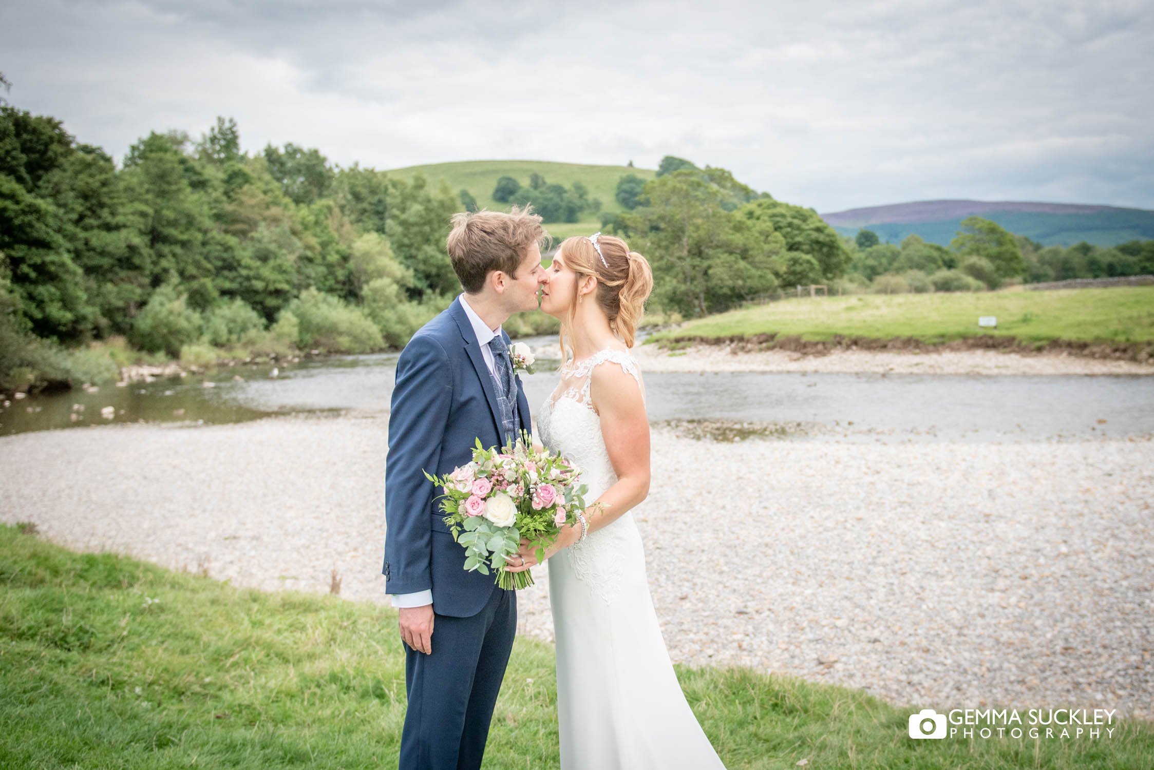 bride and groom kissing with burnsall river behind them