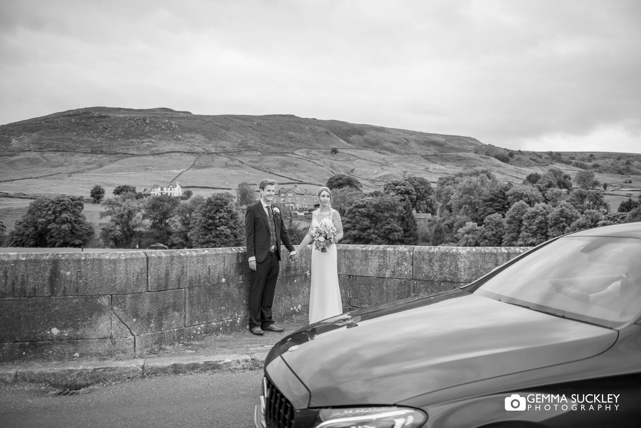 bride and groom on burnsall bridge as a car passes