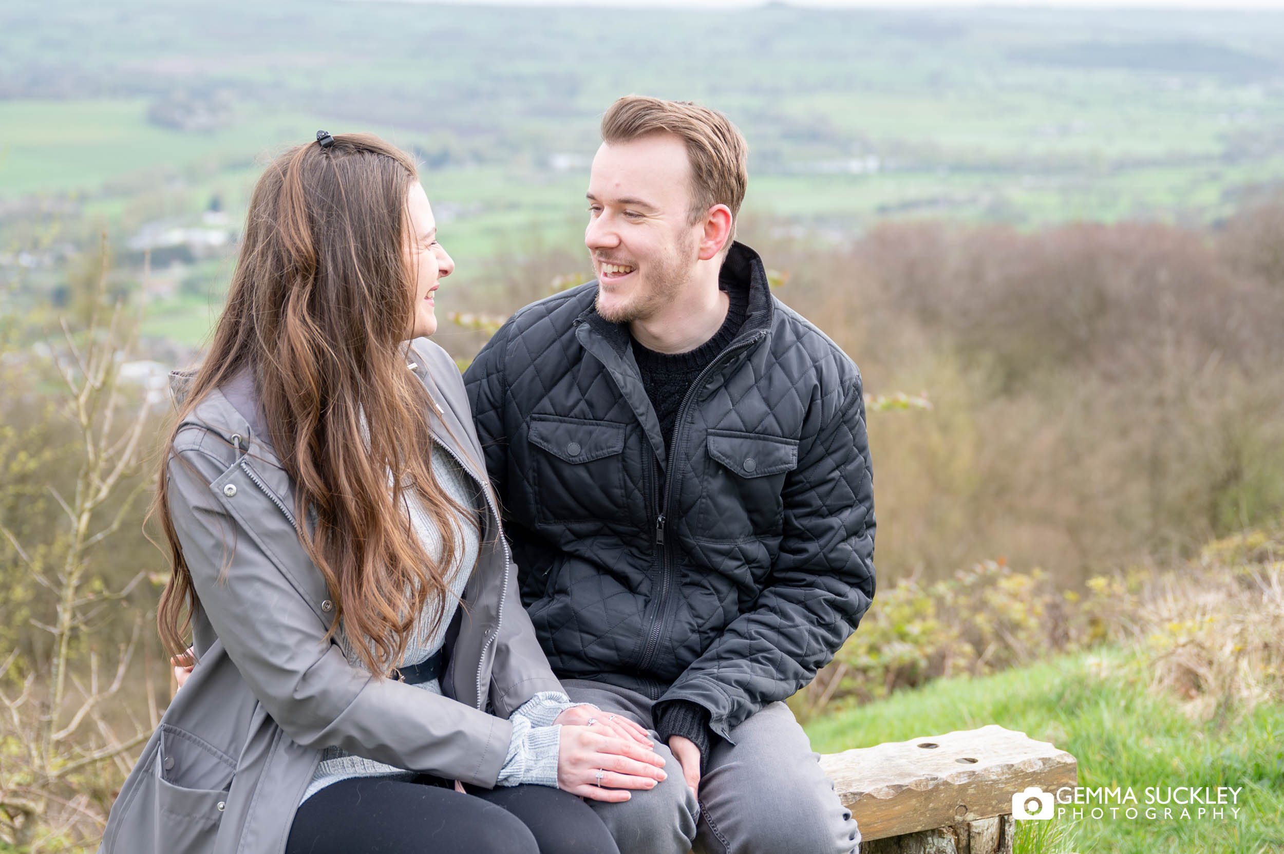 engaged couple sitting on a bench on otley chevin with the view of otley town behind them