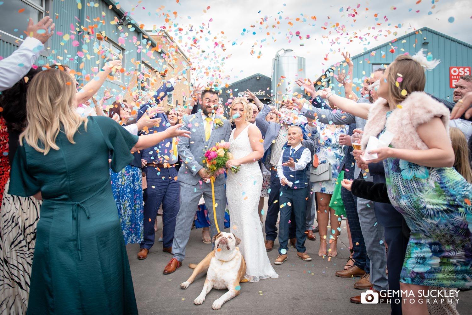 Wedding guests throwing colourful confetti at newly-weds and their dog
