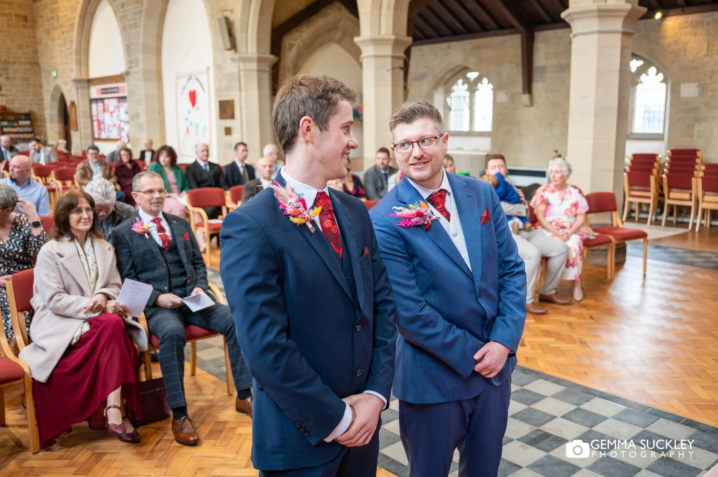 the groom and bestman talking at the top of the church aisle