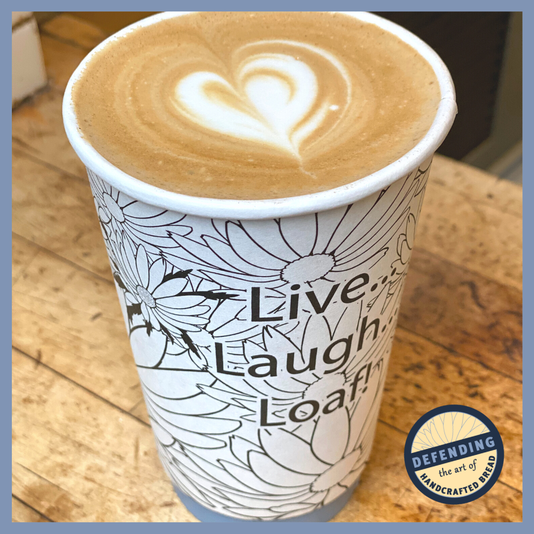 An espresso latte made at Grandaisy Bakery using Illy brand espresso sitting on rustic wooden counter. The latte is in a paper cup that has pattern of black and white daisies, and the words, "Live, Laugh, Loaf!" and latte-art in the shape of a heart