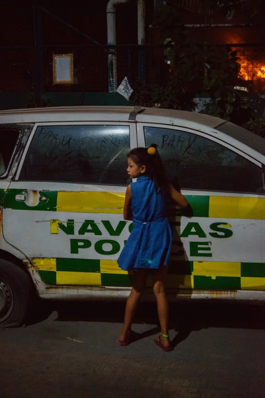 A girl writes on the window of a parked police car as a fire burns in the background. NAVOTAS, MANILA. 2018.