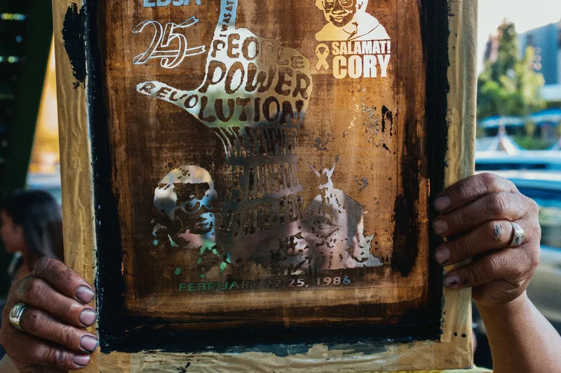A street vendor holds up a mold for a screen print design that he is selling on t-shirts to commemorate the 25th anniversary of the EDSA Revolution. The People Power Revolution (also known as the EDSA Revolution and the Philippine Revolution of 1986 or simply EDSA 1986) was a series of popular demonstrations in the Philippines, mostly in the capital city of Manila from February 22–25, 1986. There was a sustained campaign of civil resistance against regime violence and alleged electoral fraud. The nonviolent revolution led to the departure of dictator Ferdinand Marcos, the end of his 21-year totalitarian rule, and the restoration of democracy in the Philippines. QUEZON CITY, MANILA. 2011.