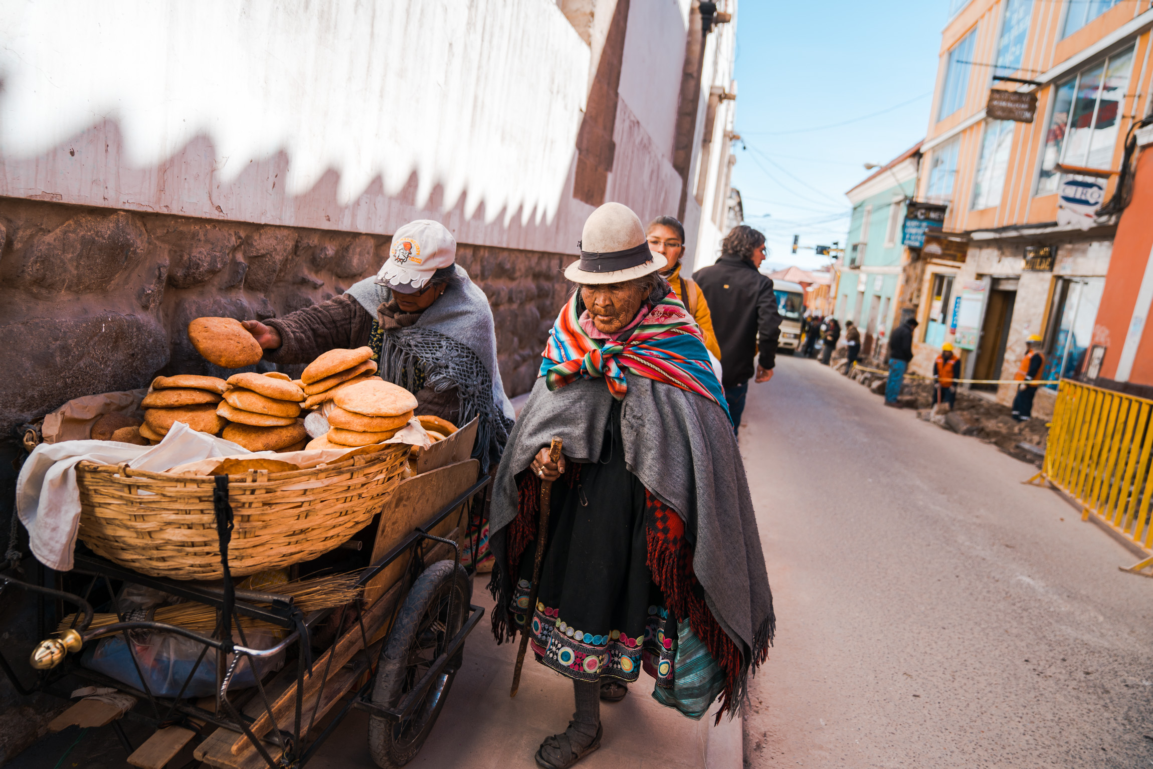 Bolivian woman walks past someone selling traditional break on the street side. Potosi, Bolivia. 2018