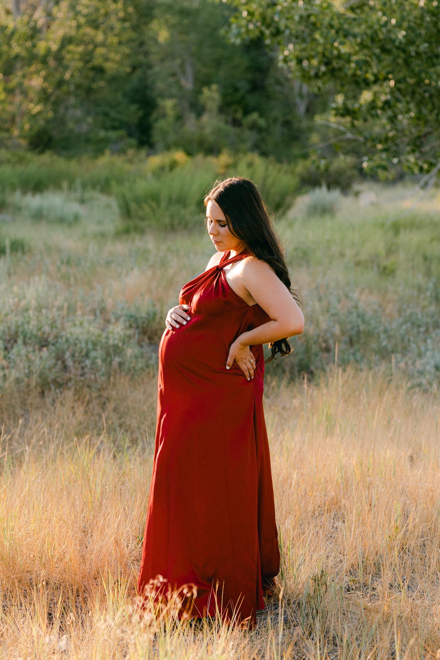Maternity session in the meadows, mom to be wearing a red maternity gown and standing in the meadow with golden light hitting her face